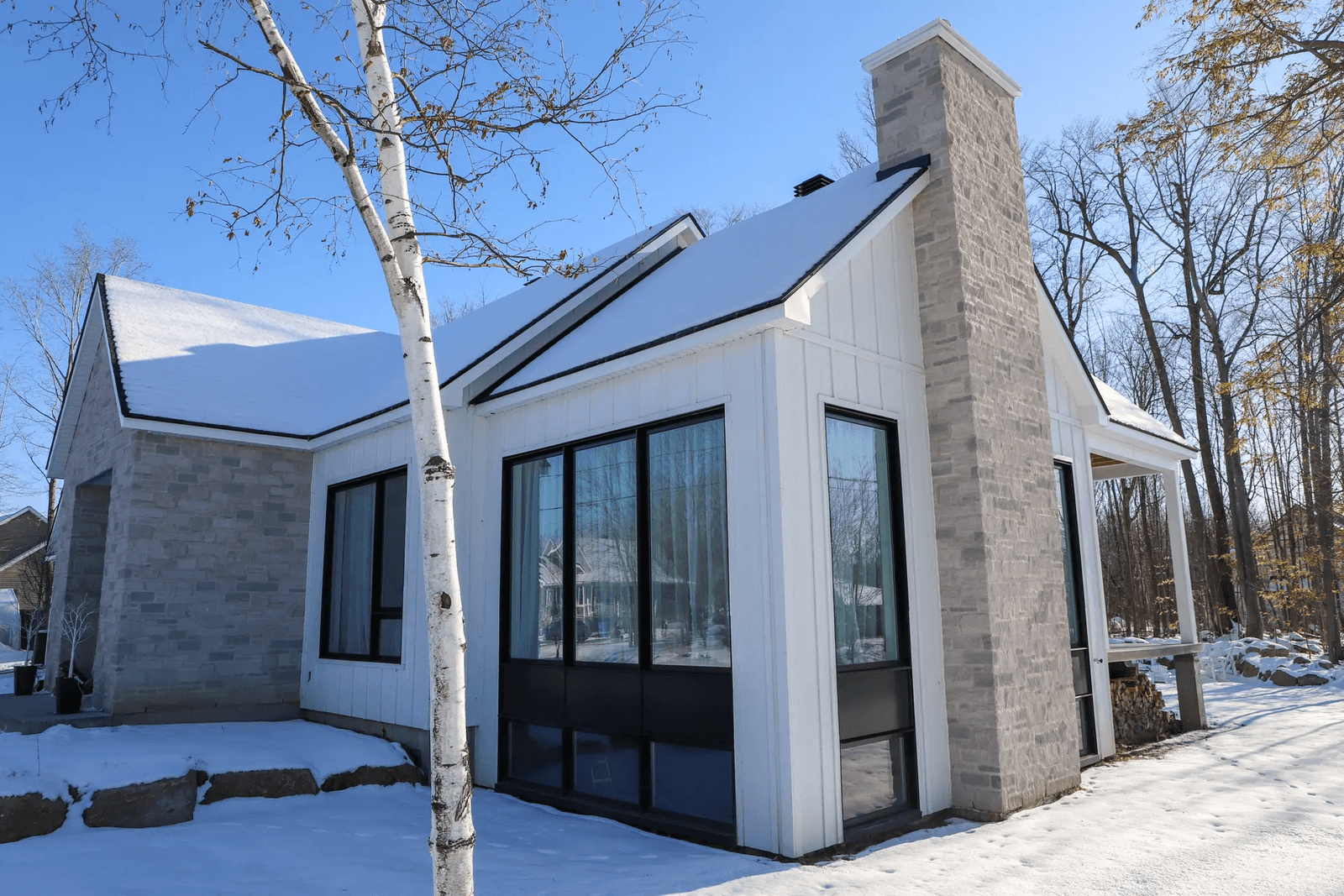 Modern house in winter with white wood and stone siding, large windows, and stone chimney in a wooded setting.