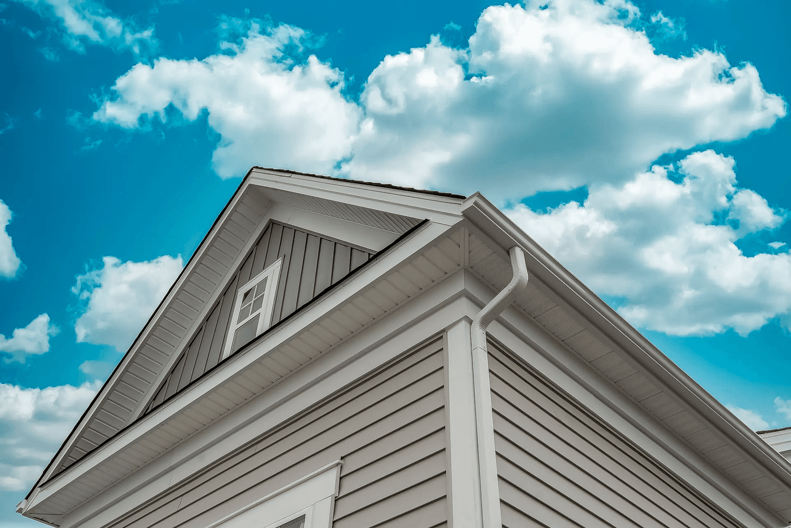 House with beige vinyl siding, white windows, and gutters under a cloudy blue sky.