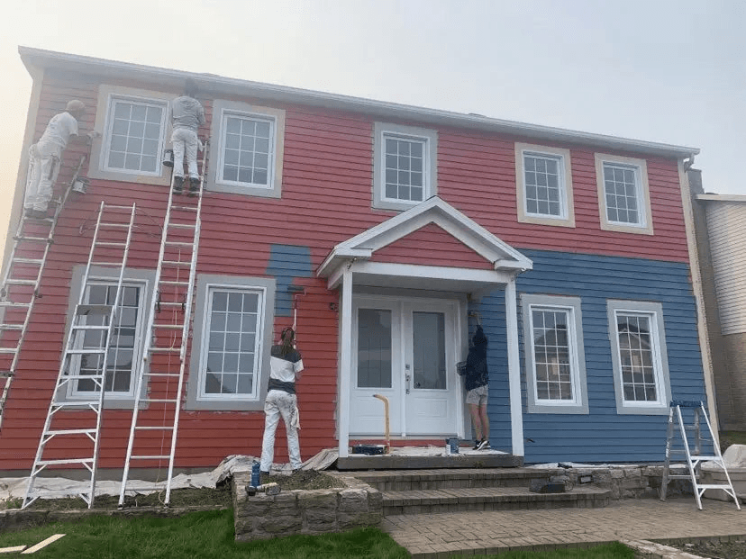 Workers repainting the exterior siding of a two-story house using ladders, showing a facade transition from red to dark blue.