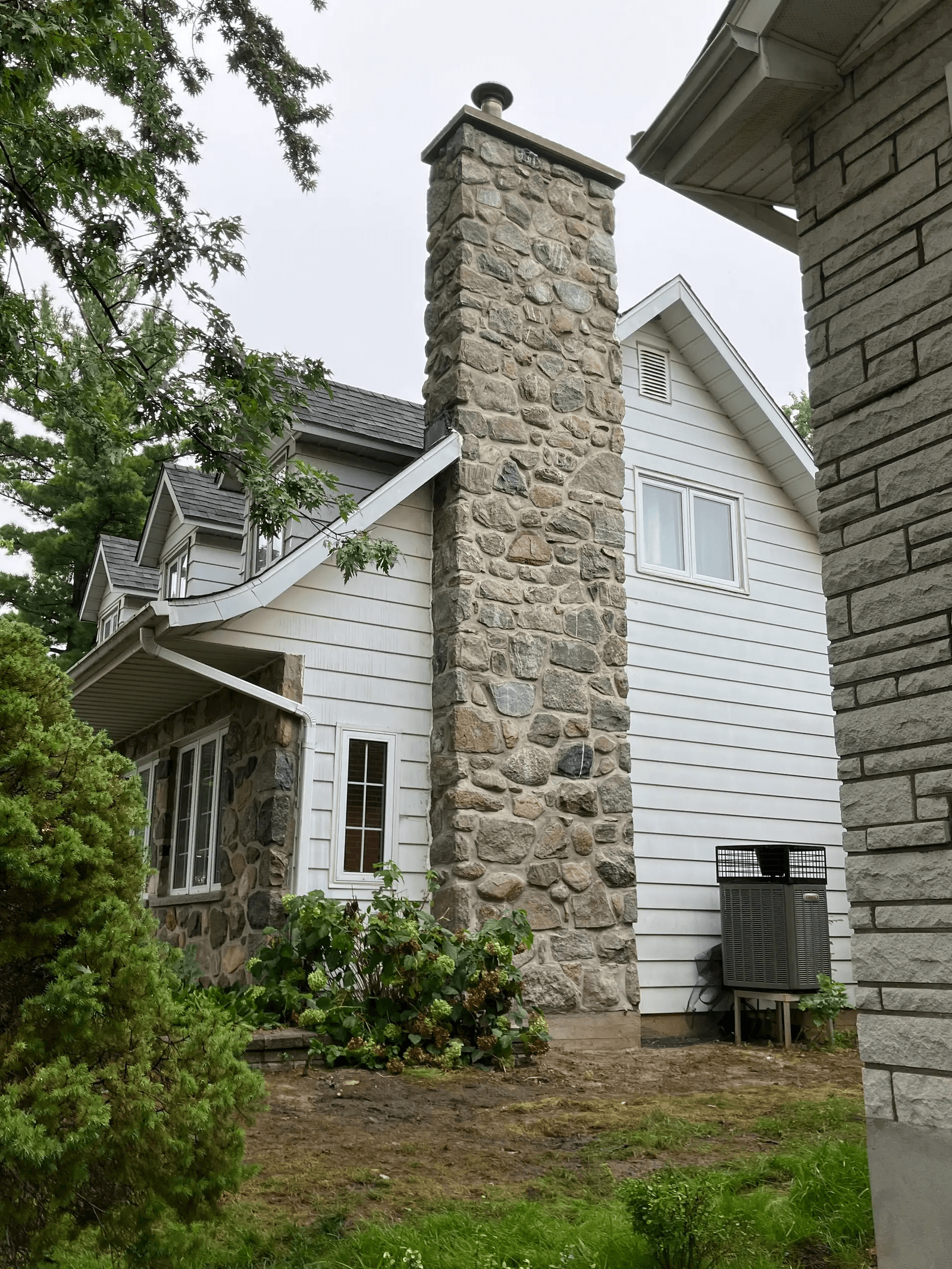 Large exterior natural stone chimney on a residential house featuring white siding, a shingle roof and lush green landscaping