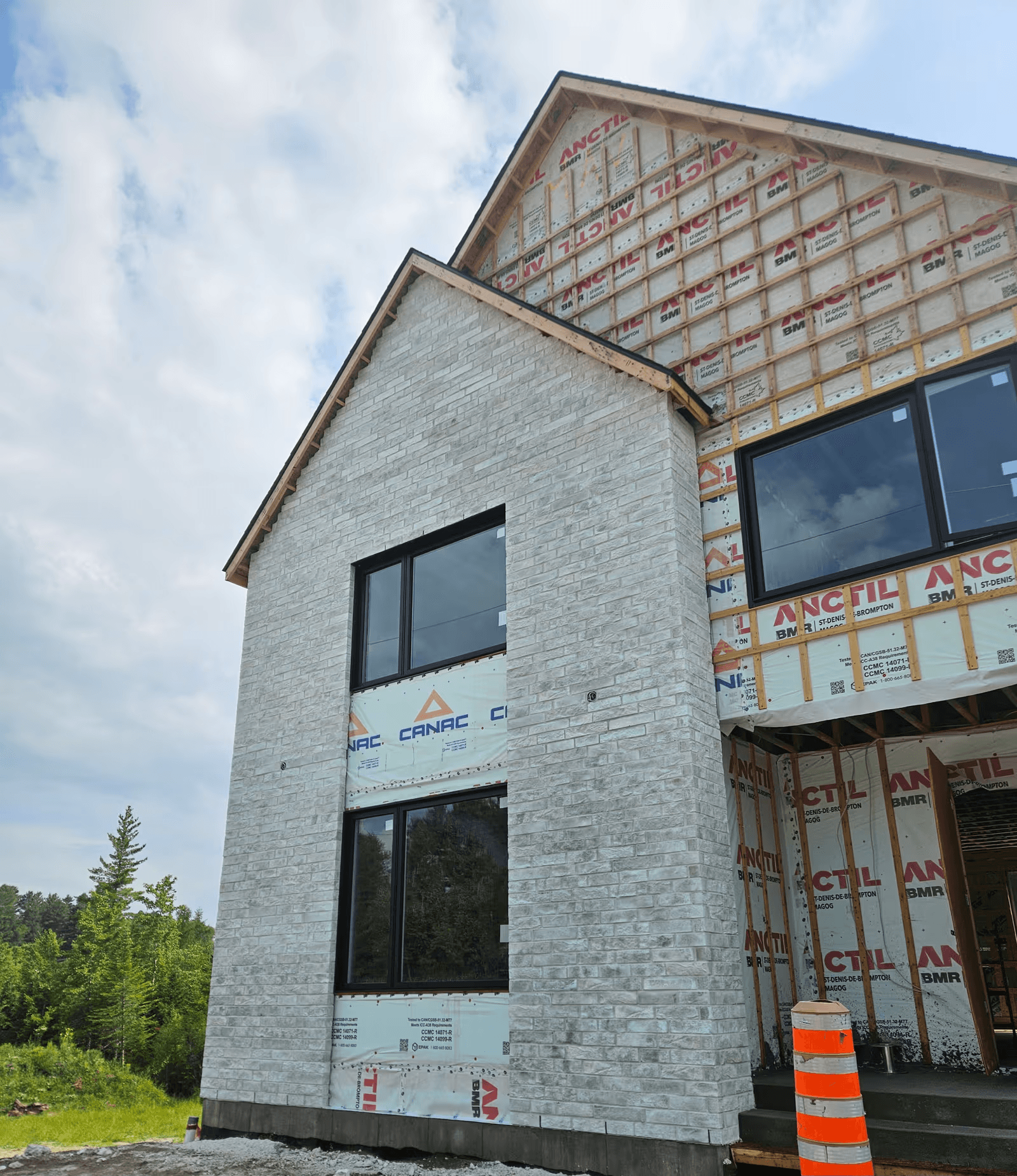 New home under construction with partially installed white brick veneer, large black windows, exterior weather barrier and exposed wood furring