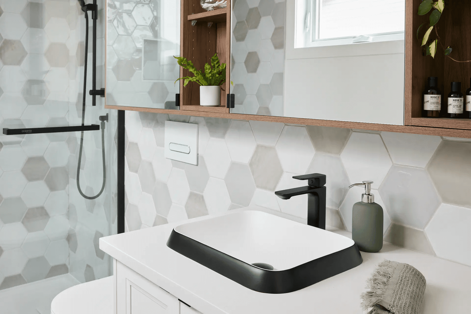 Modern bathroom featuring white, grey, and beige hexagonal wall tiles, a glass-enclosed shower with black fixtures, a rectangular black-and-white vessel sink, white countertop, wooden vanity, and wall-mounted cabinets with decorative storage.