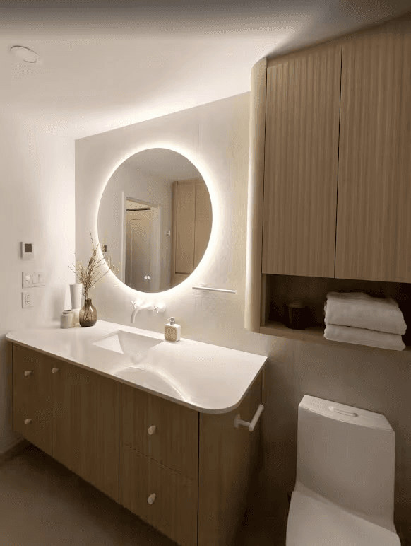 Modern bathroom featuring a floating natural wood vanity, an integrated white sink, a backlit round mirror, and minimalist cabinetry.