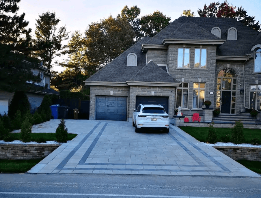 Residential grey interlocking paver driveway with dark decorative patterns, double garage, landscaped retaining walls and stone house