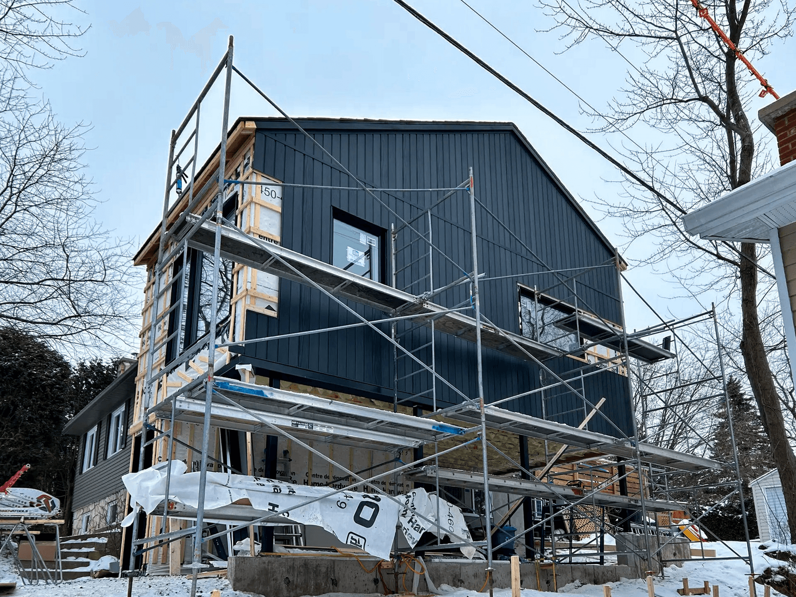 Residential home extension under construction with scaffolding, black metal exterior siding, new windows and timber framing on a concrete foundation.