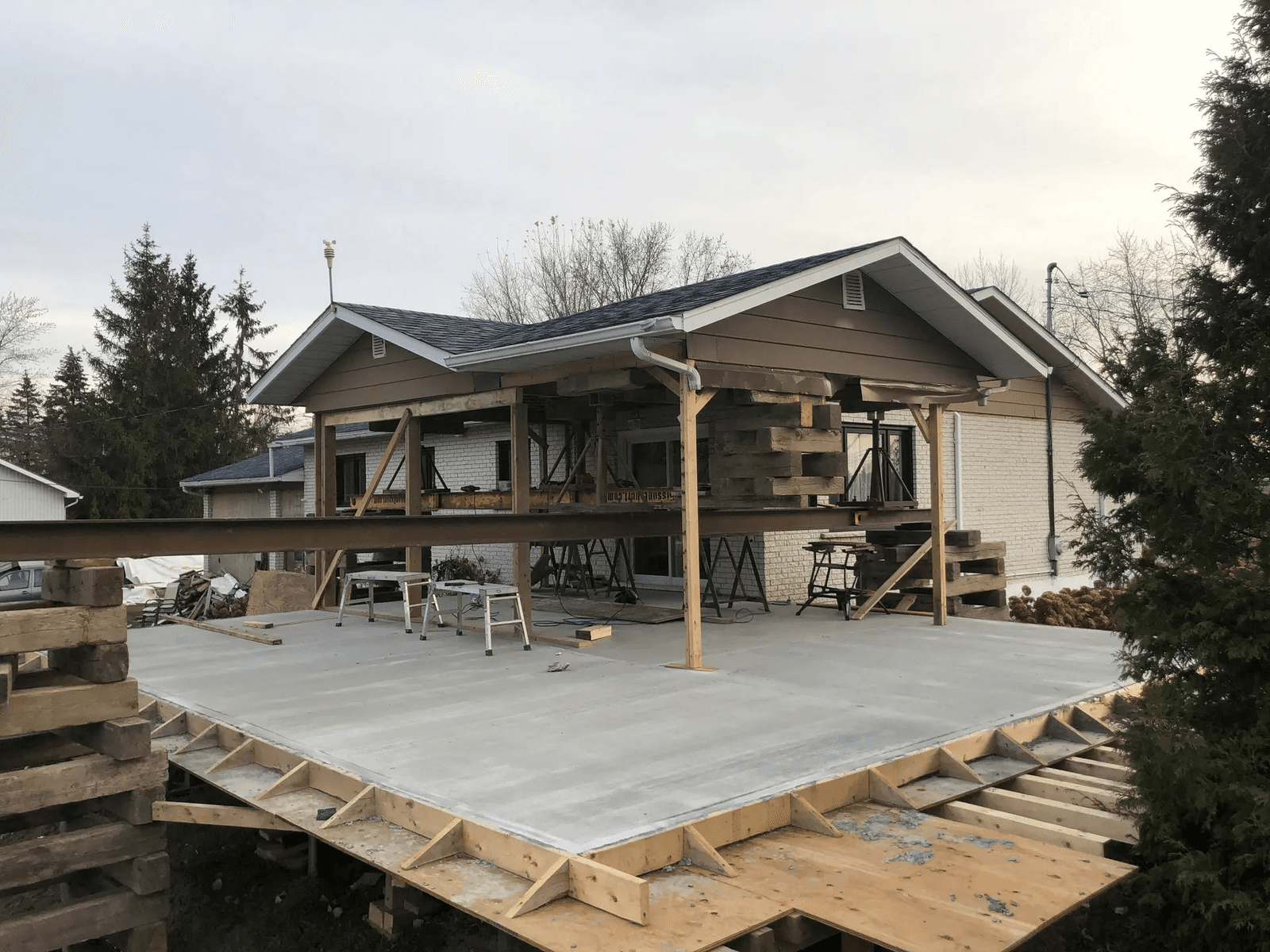 Residential home extension under construction with concrete slab, temporary structural supports, steel beams and timber framing at the rear of a house.