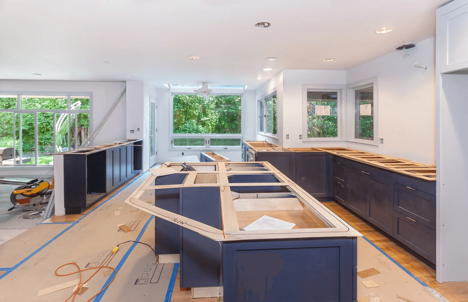 Residential kitchen under renovation with dark blue cabinets being installed, a large central island, protected flooring, recessed lighting, and large windows overlooking the backyard