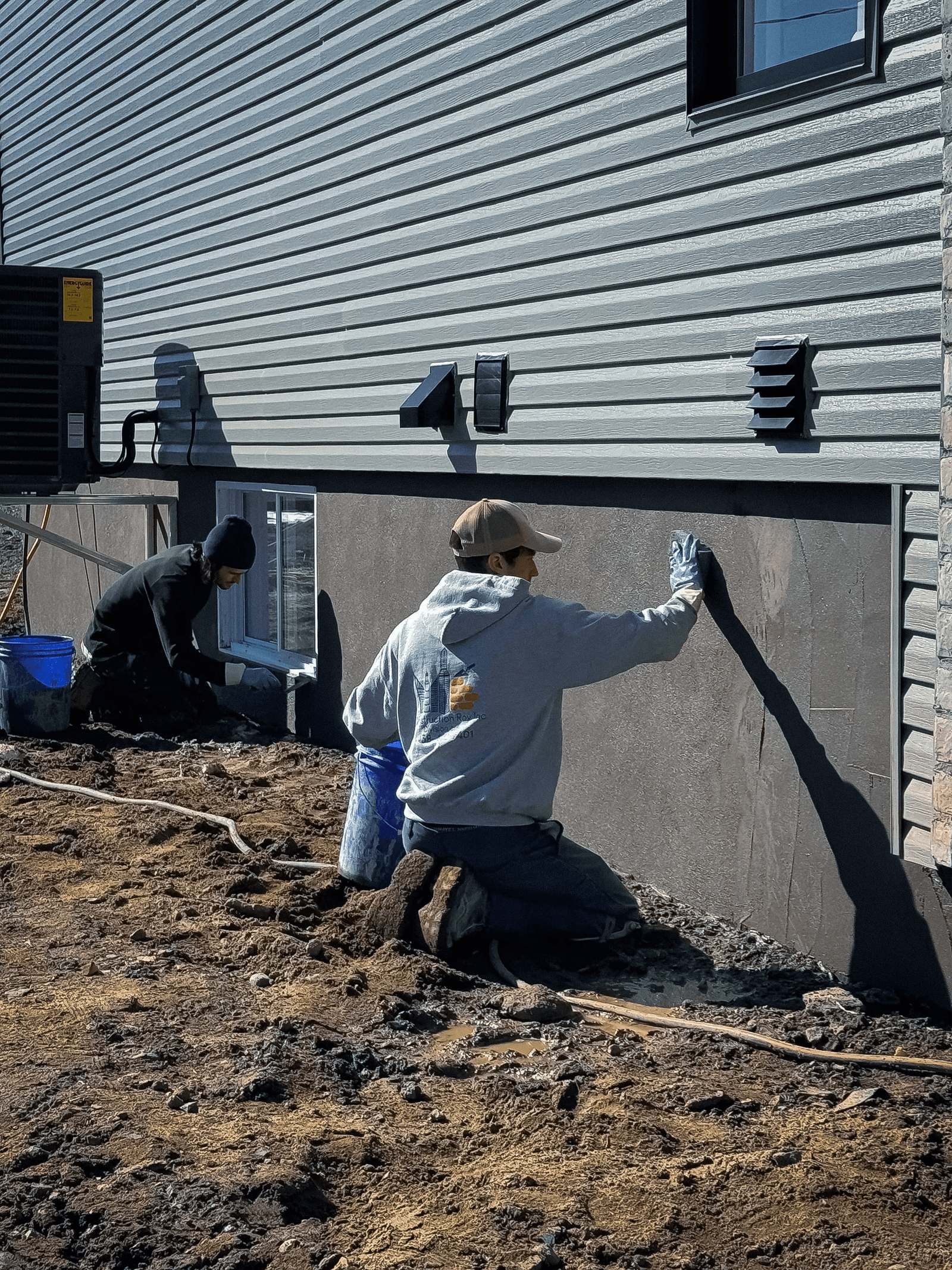 Workers applying a coating to a house foundation as part of crack repair and waterproofing work.