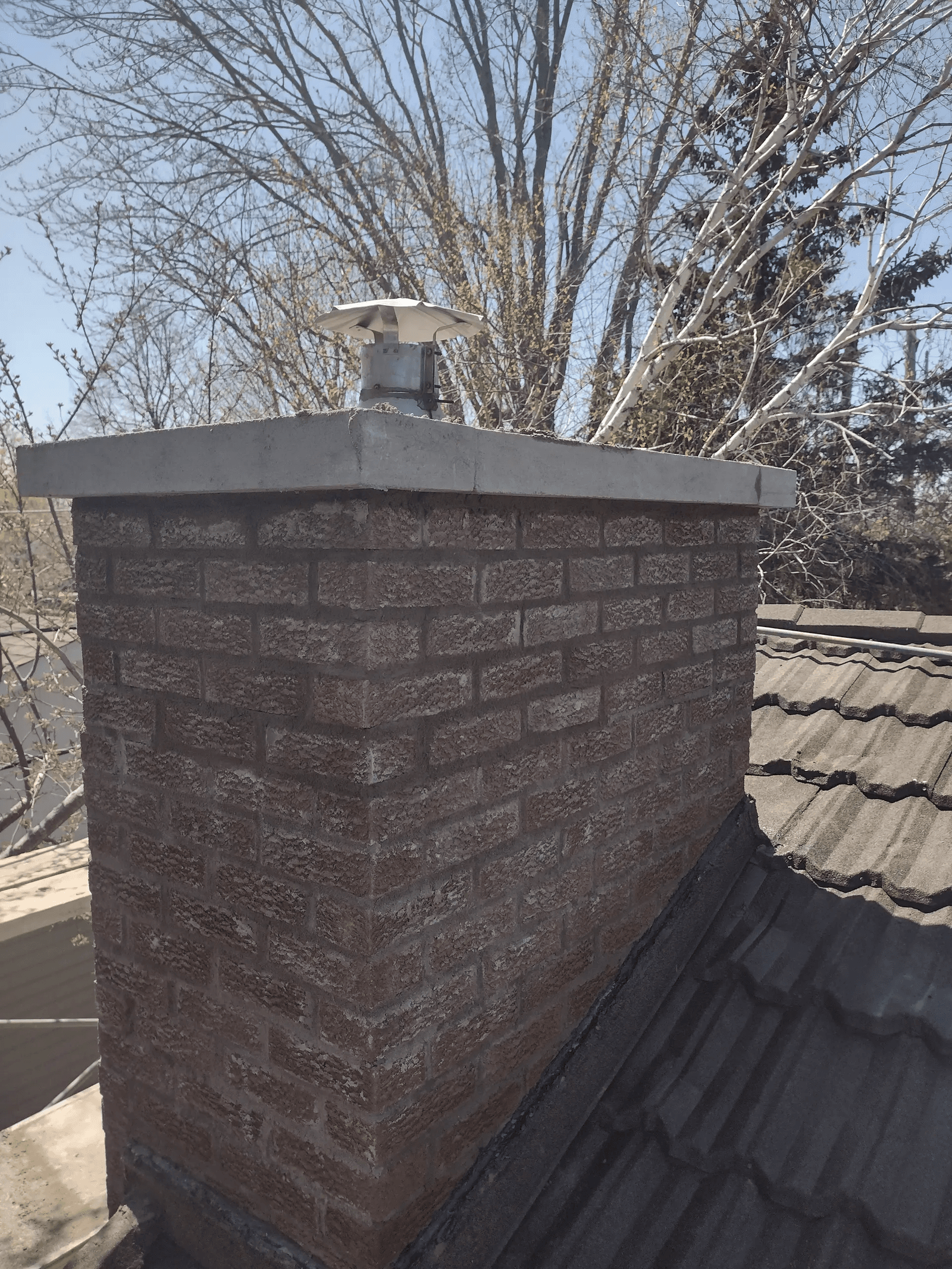 Exterior red brick chimney with a concrete crown and metal chimney cap, installed on a residential roof with grey shingles