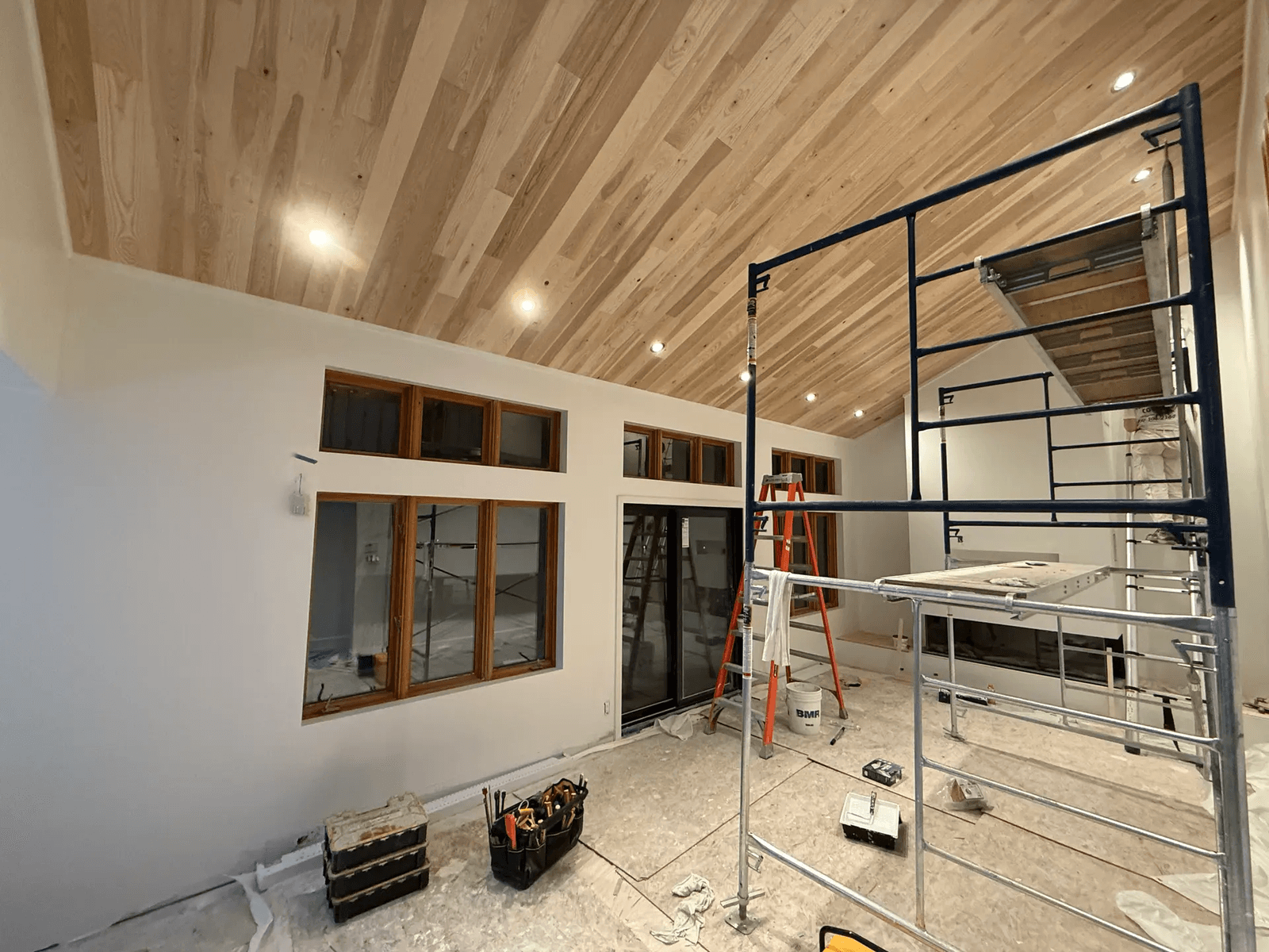 &nbsp;Interior renovation in progress with a cathedral ceiling clad in wood slats, recessed lighting, large windows, glass doors, scaffolding, and construction tools