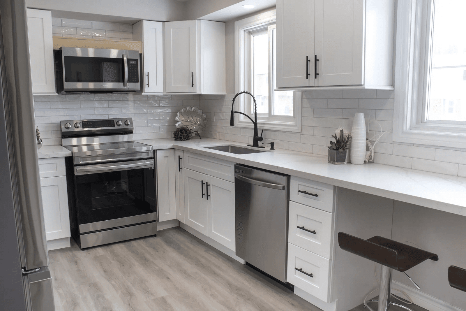Bright modern kitchen with white cabinets, light countertops, ceramic tile backsplash, stainless steel appliances, sink with black faucet and breakfast bar with stools.