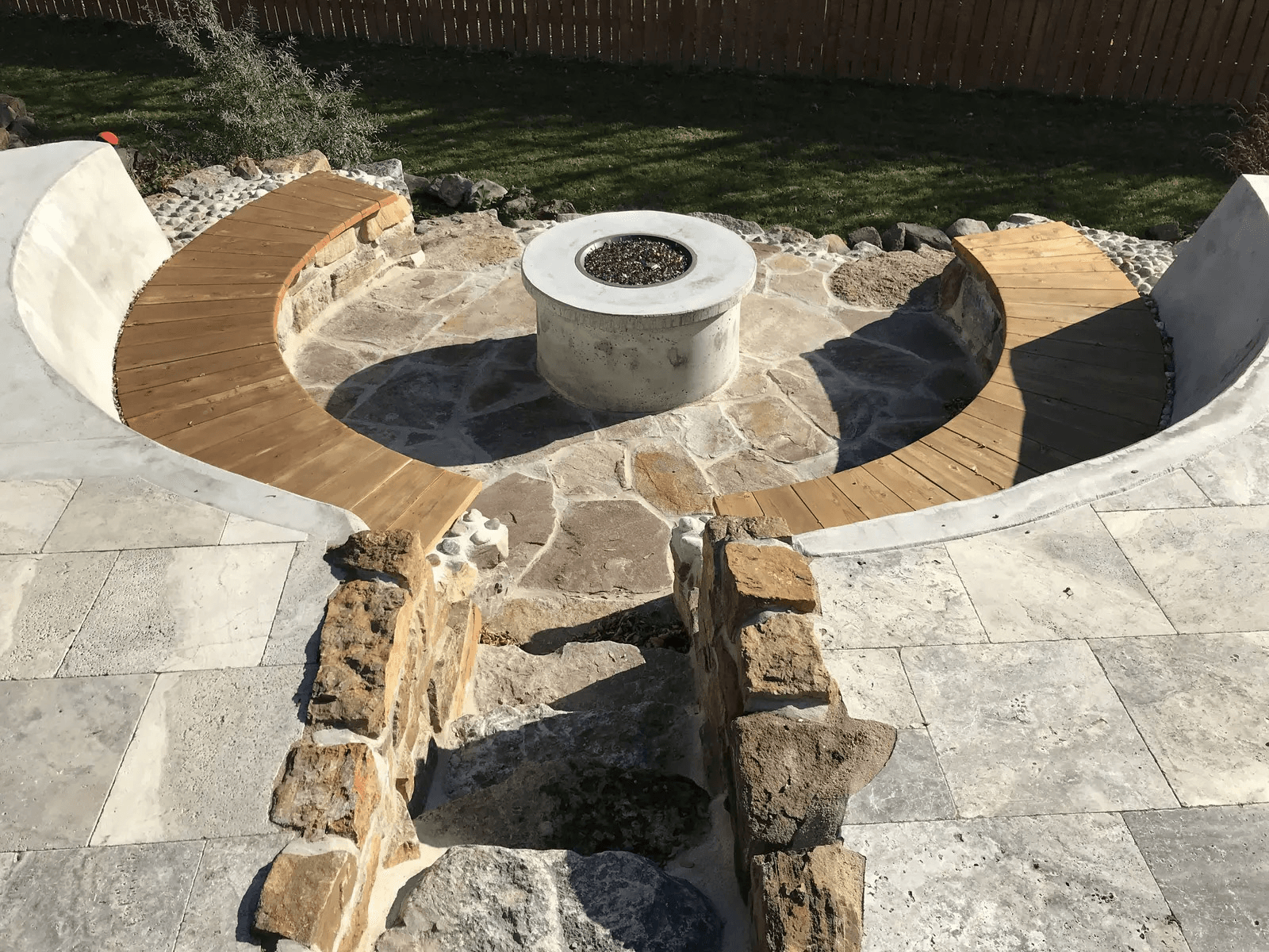 Circular outdoor landscaping with central concrete fire pit, curved wooden benches, natural stone patio, masonry walls and landscaped lawn.