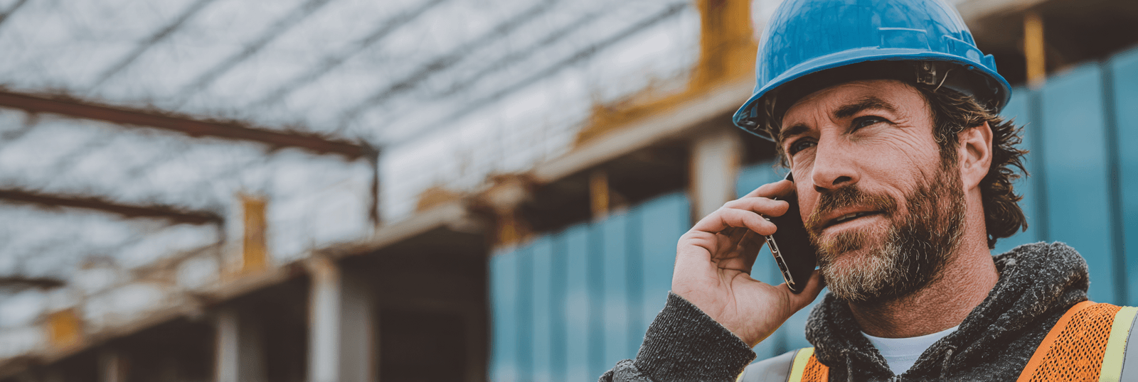 Construction professional wearing a blue hard hat and high-visibility vest talking on a phone at a building site under construction