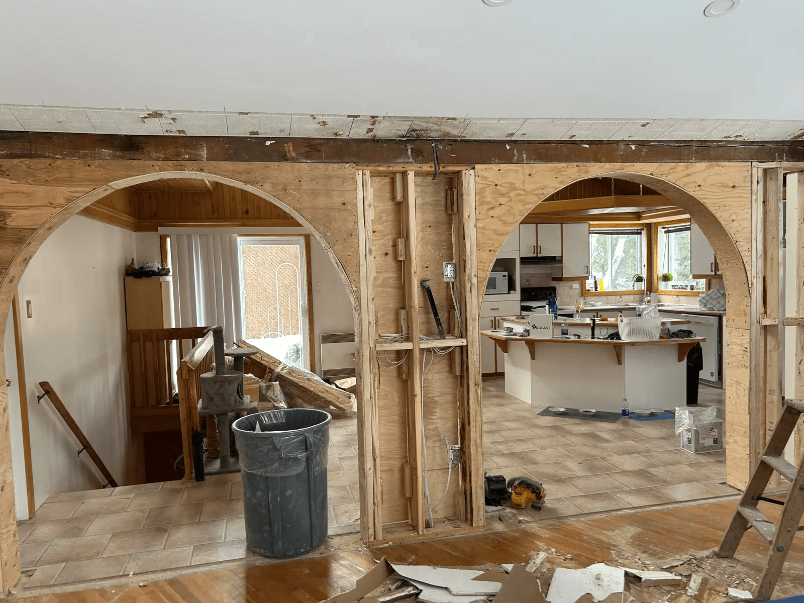 Residential interior renovation in progress with an opened load-bearing wall, large wood arches, exposed framing, electrical wiring, and kitchen visible in the background