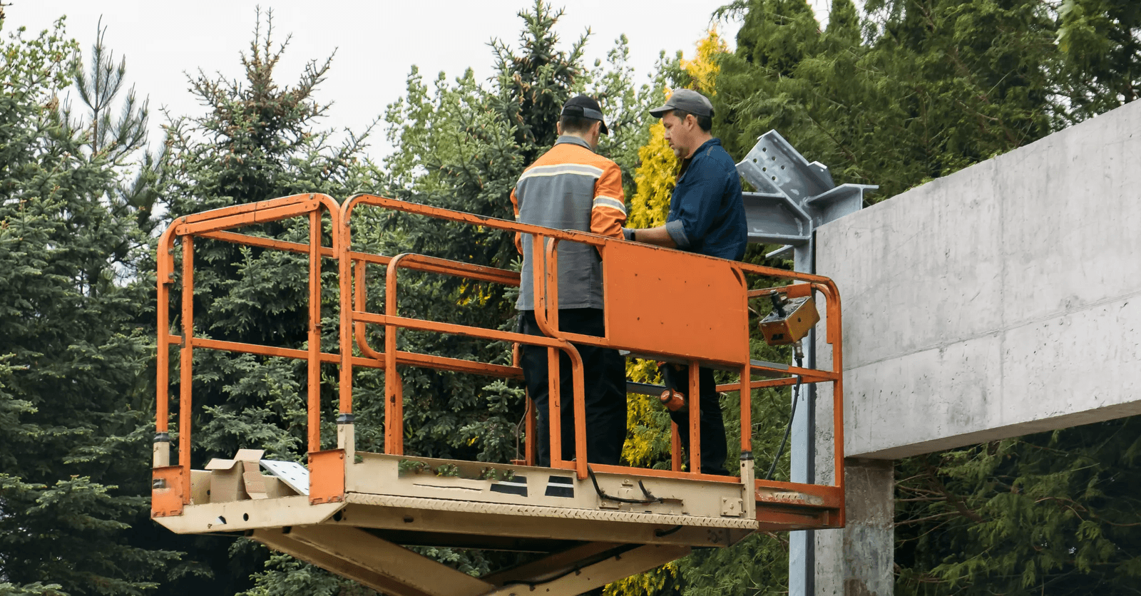 Two workers on an orange scissor lift carrying out installation and assembly work on a metal structure attached to an exterior concrete wall, on a construction site surrounded by coniferous trees.