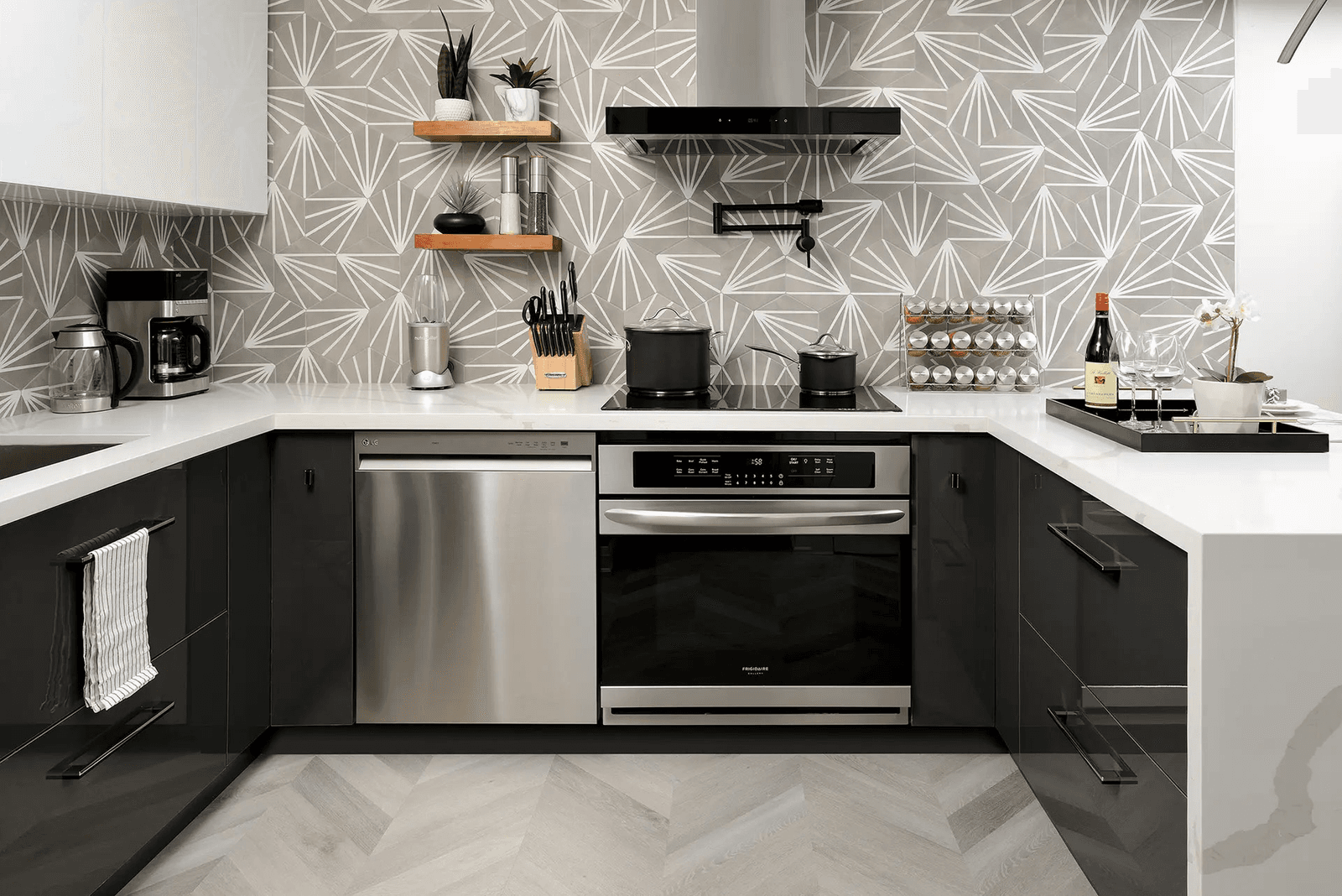 Contemporary kitchen with black cabinets, white countertops, geometric backsplash, stainless steel appliances, black range hood and chevron-pattern flooring.