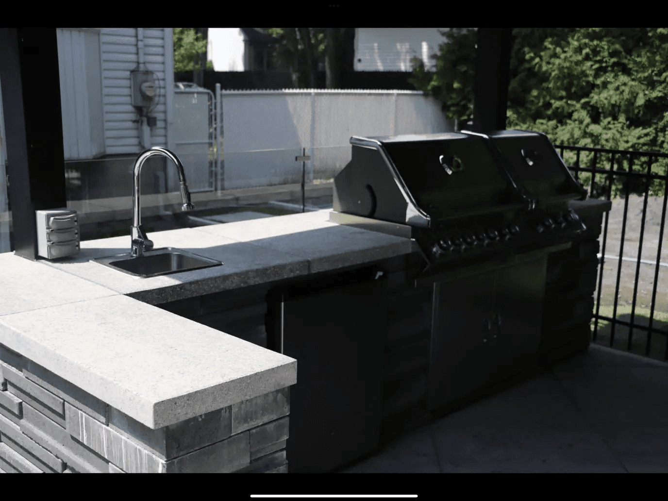 Outdoor kitchen with a built-in barbecue, sink, stone countertops and integrated storage under a covered structure.