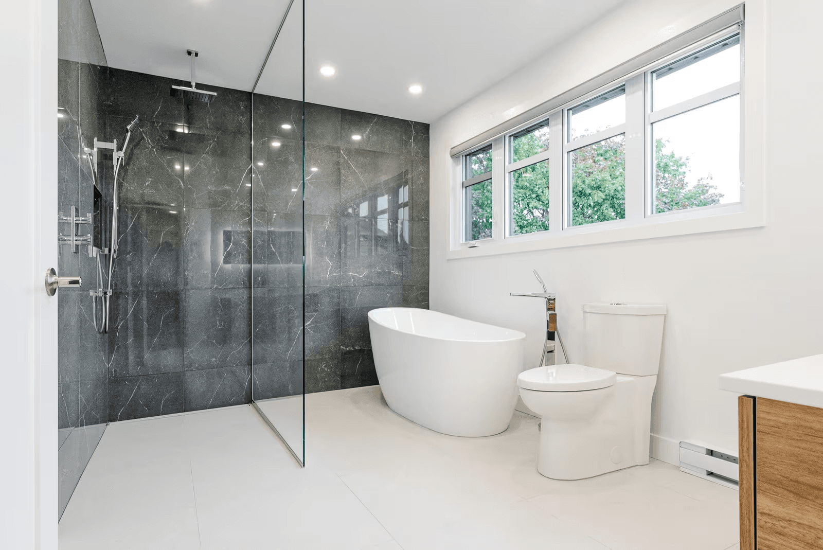 Modern bathroom with a glass walk-in shower, large anthracite stone-look tiles, white freestanding bathtub and toilet beneath a wide window