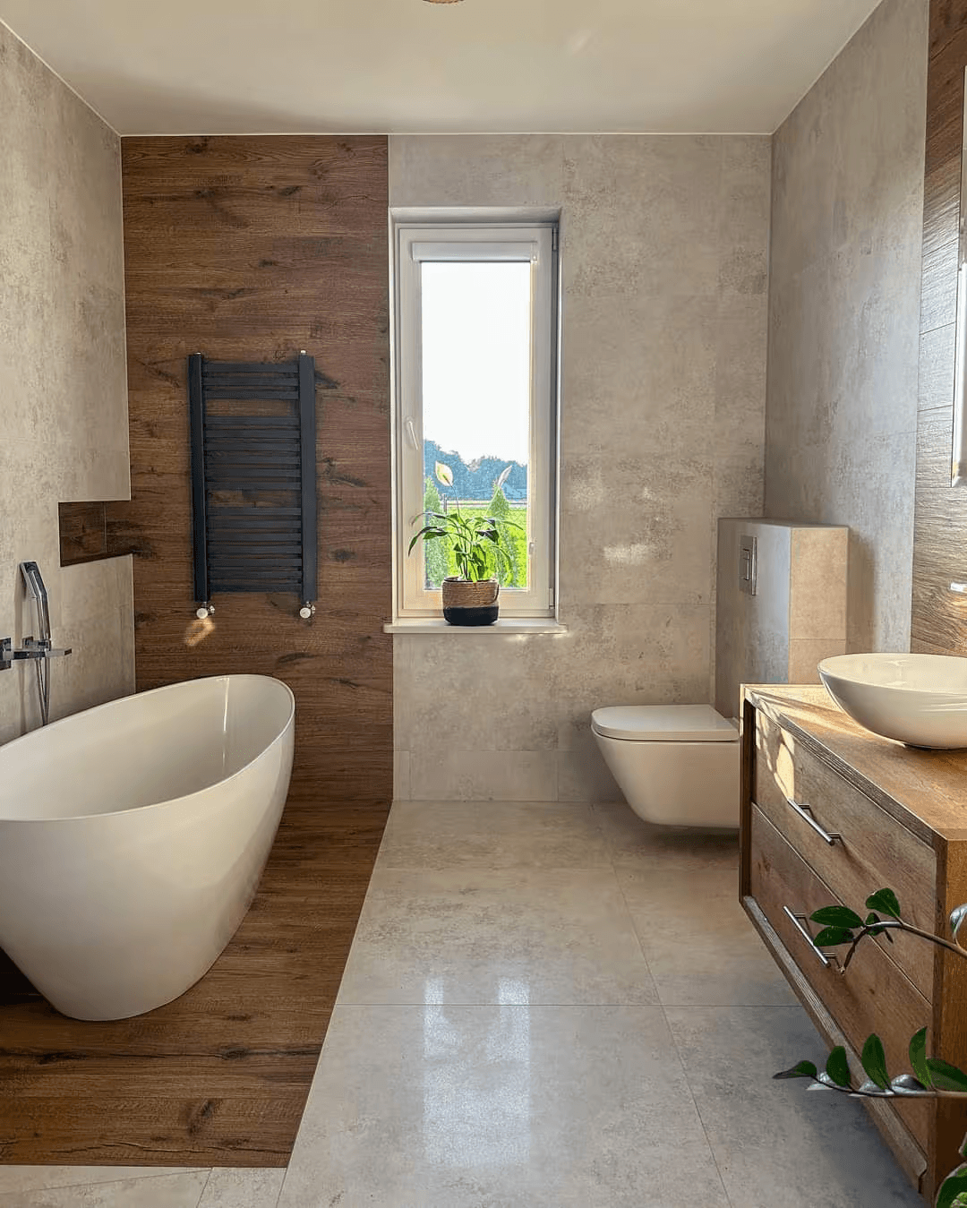 Contemporary bathroom featuring a white freestanding bathtub, brown wood-look wall tiles, large beige stone-look ceramic tiles, wall-mounted toilet, natural wood vanity, white vessel sink, black towel warmer, and bright window.
