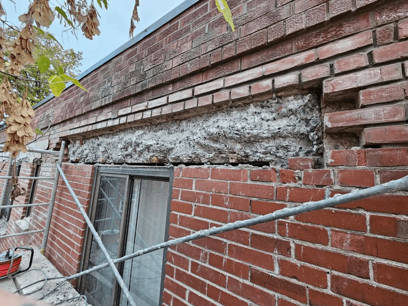 Exterior brick wall under repair with masonry removed above a window, exposed concrete and scaffolding installed along the building