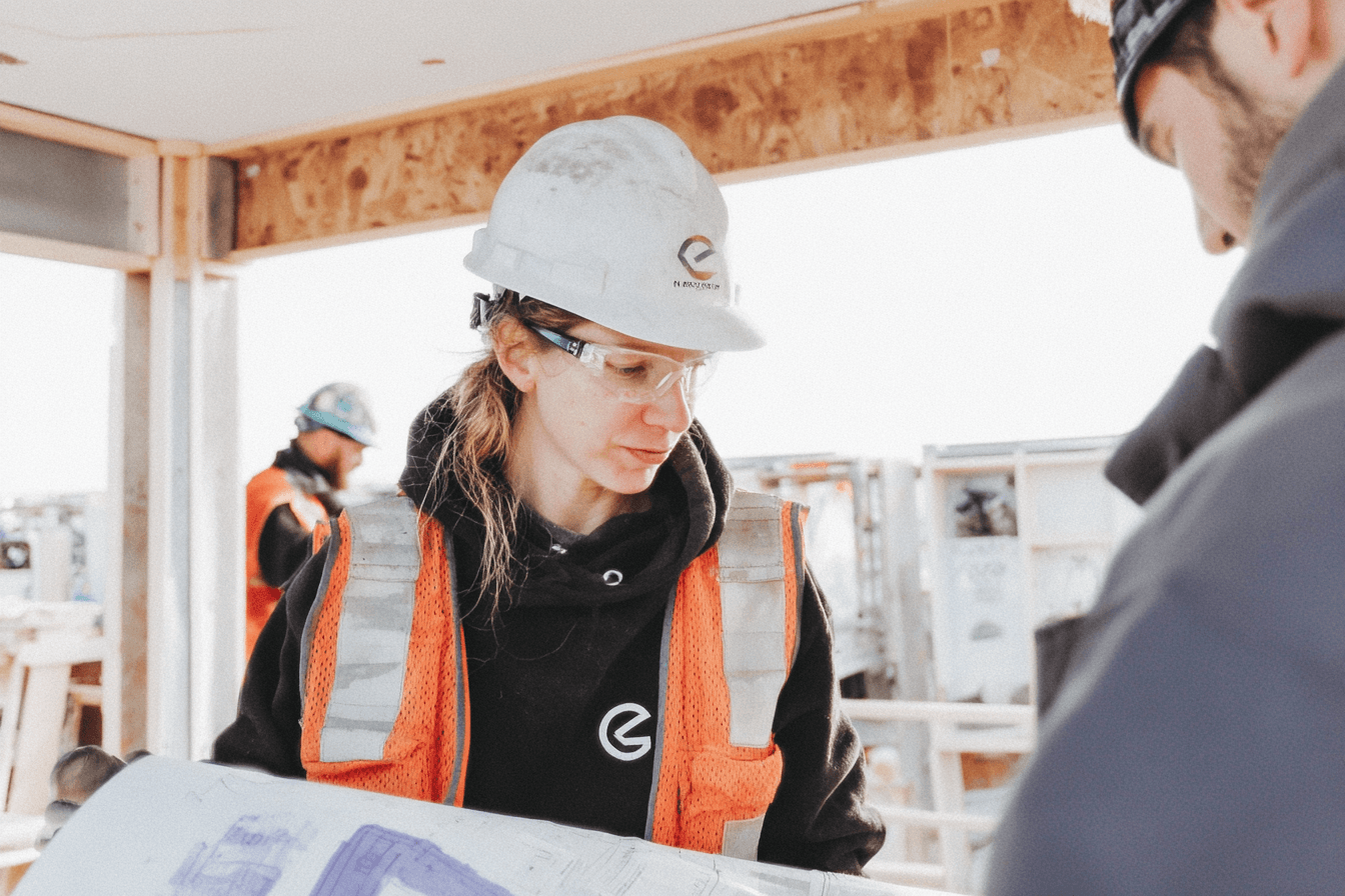 Construction professional wearing a white hard hat, safety glasses and a high-visibility vest reviewing plans on an indoor renovation site