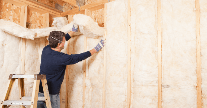Worker installing batt insulation between wood studs inside a building under construction.