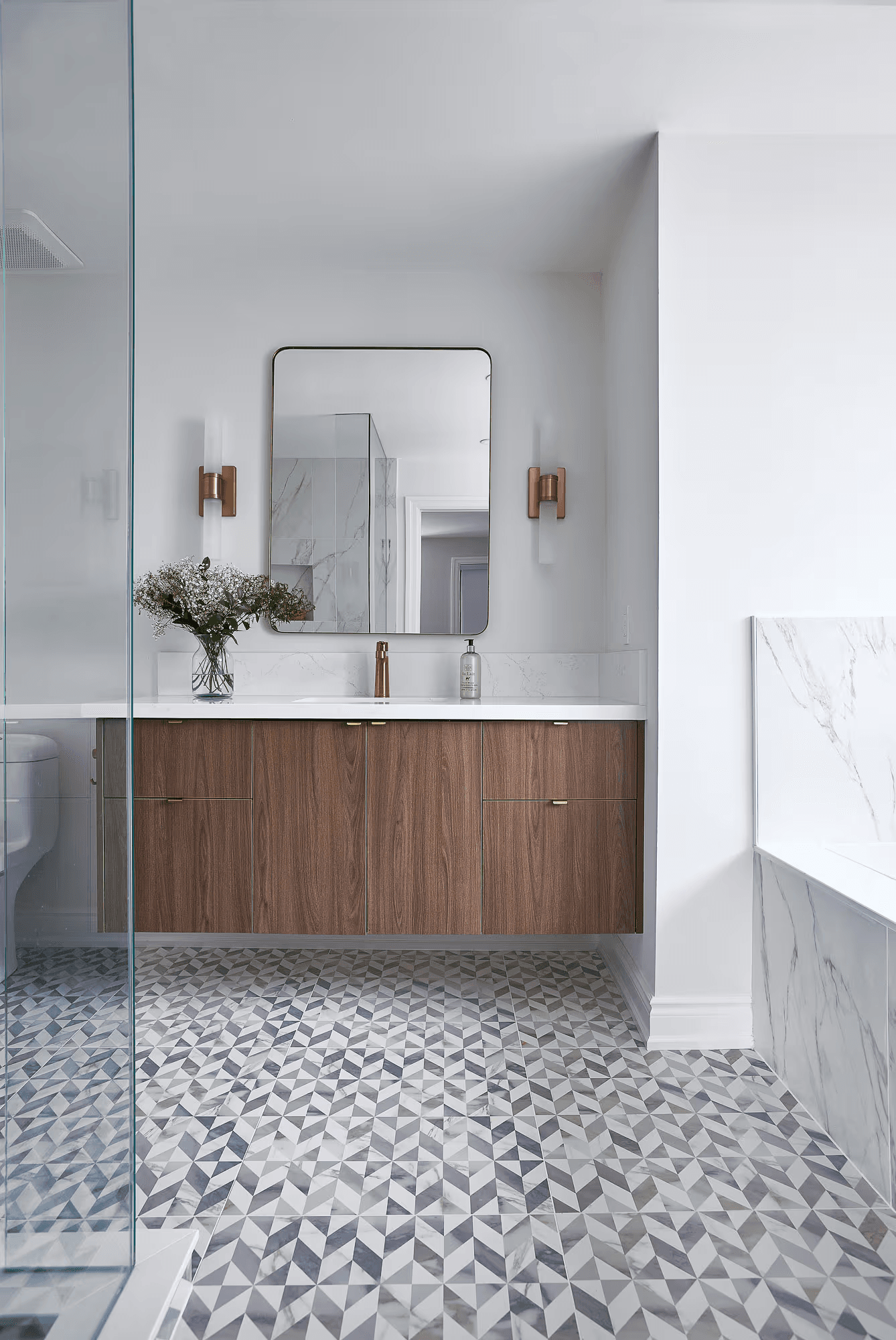 Upscale contemporary bathroom featuring a brown wooden vanity, white countertop, gold fixtures, patterned ceramic flooring, a glass shower, and a bathtub.