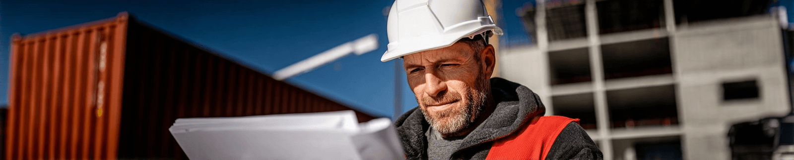 Construction contractor wearing a white hard hat and red safety vest reviewing plans at an outdoor commercial job site