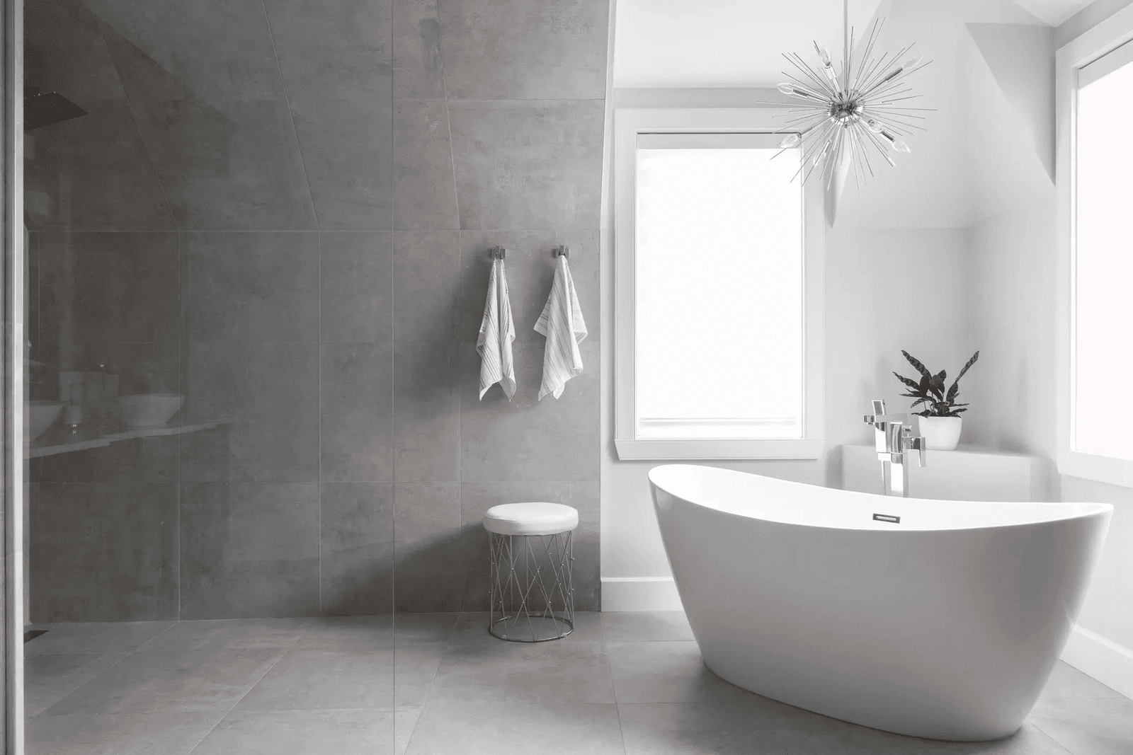 Modern luxury bathroom featuring a white freestanding bathtub, glass-enclosed walk-in shower, large grey ceramic tiles, chrome fixtures, bright windows, contemporary pendant lighting, and minimalist décor.