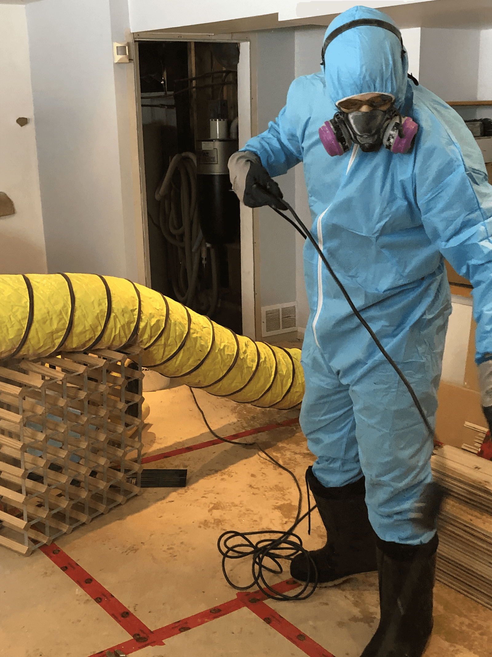 Decontamination technician wearing blue protective coveralls, a respirator and gloves during an indoor remediation job with a temporary ventilation system