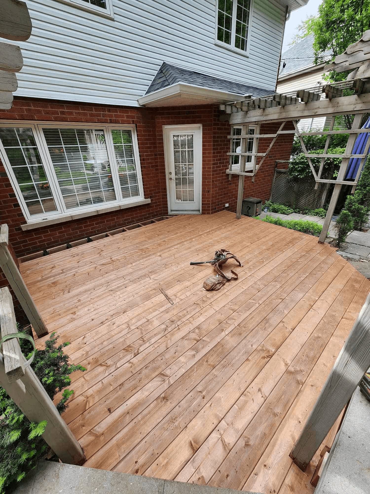 Large newly built wood deck at the back of a brick and white-sided house, with an existing pergola and access through a glass door