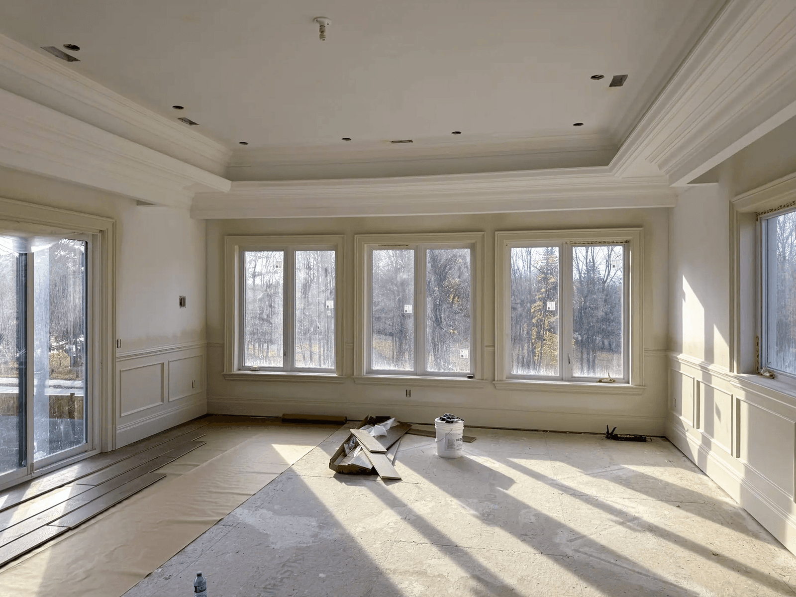 Bright room under renovation with decorative moldings, large windows, coffered ceiling, and flooring under installation.