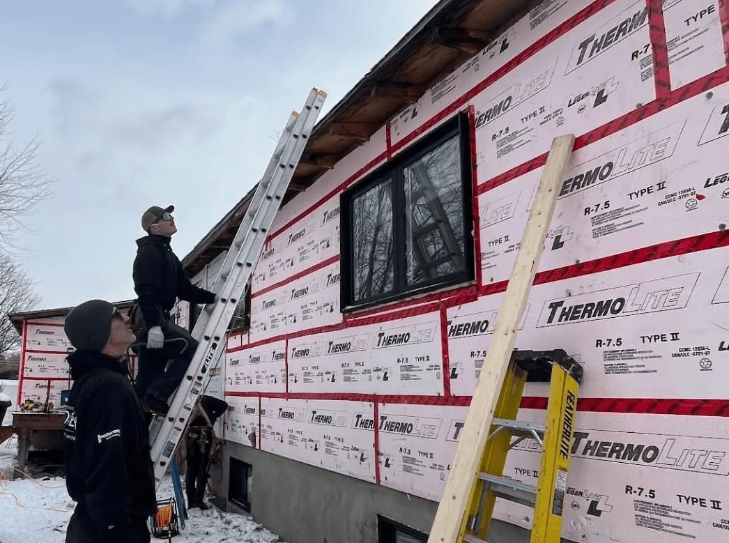Workers installing ThermoLite insulating exterior sheathing on a house under construction in winter, with ladders and construction tools.