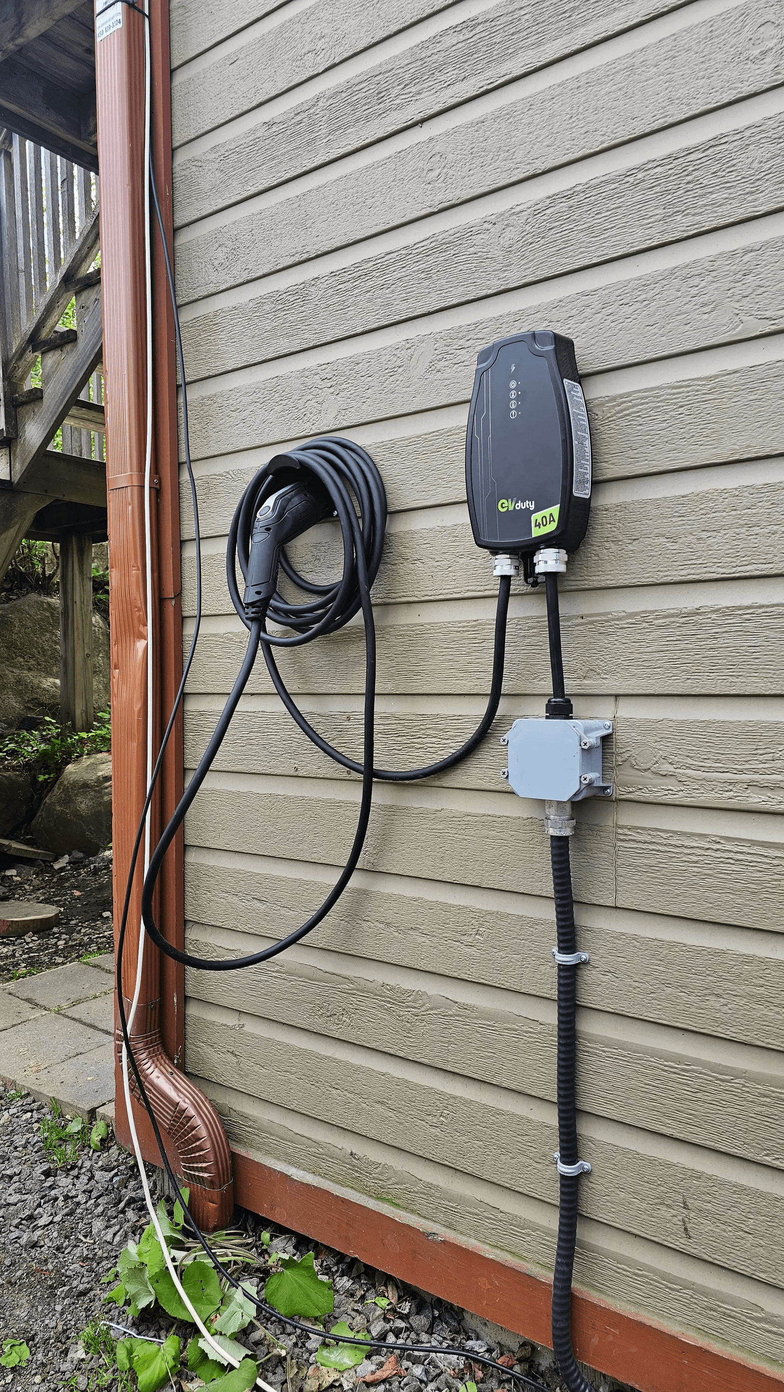 EVduty 40-amp electric vehicle charging station installed on a house's exterior siding, featuring a coiled charging cable, connector and exposed electrical connections