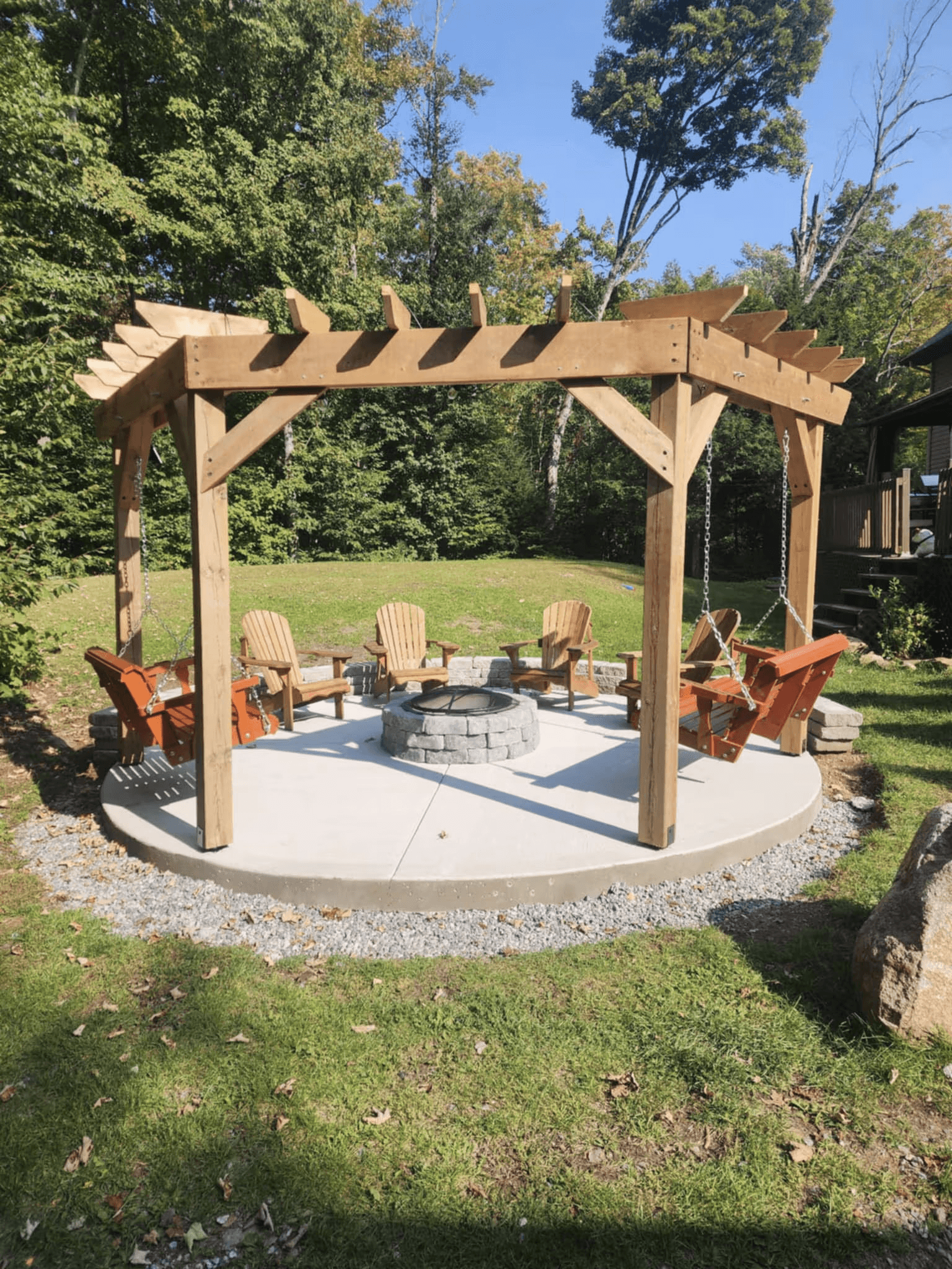 Wooden pergola built over a circular concrete patio with stone fire pit, hanging Adirondack chairs and outdoor seating area in a wooded backyard.