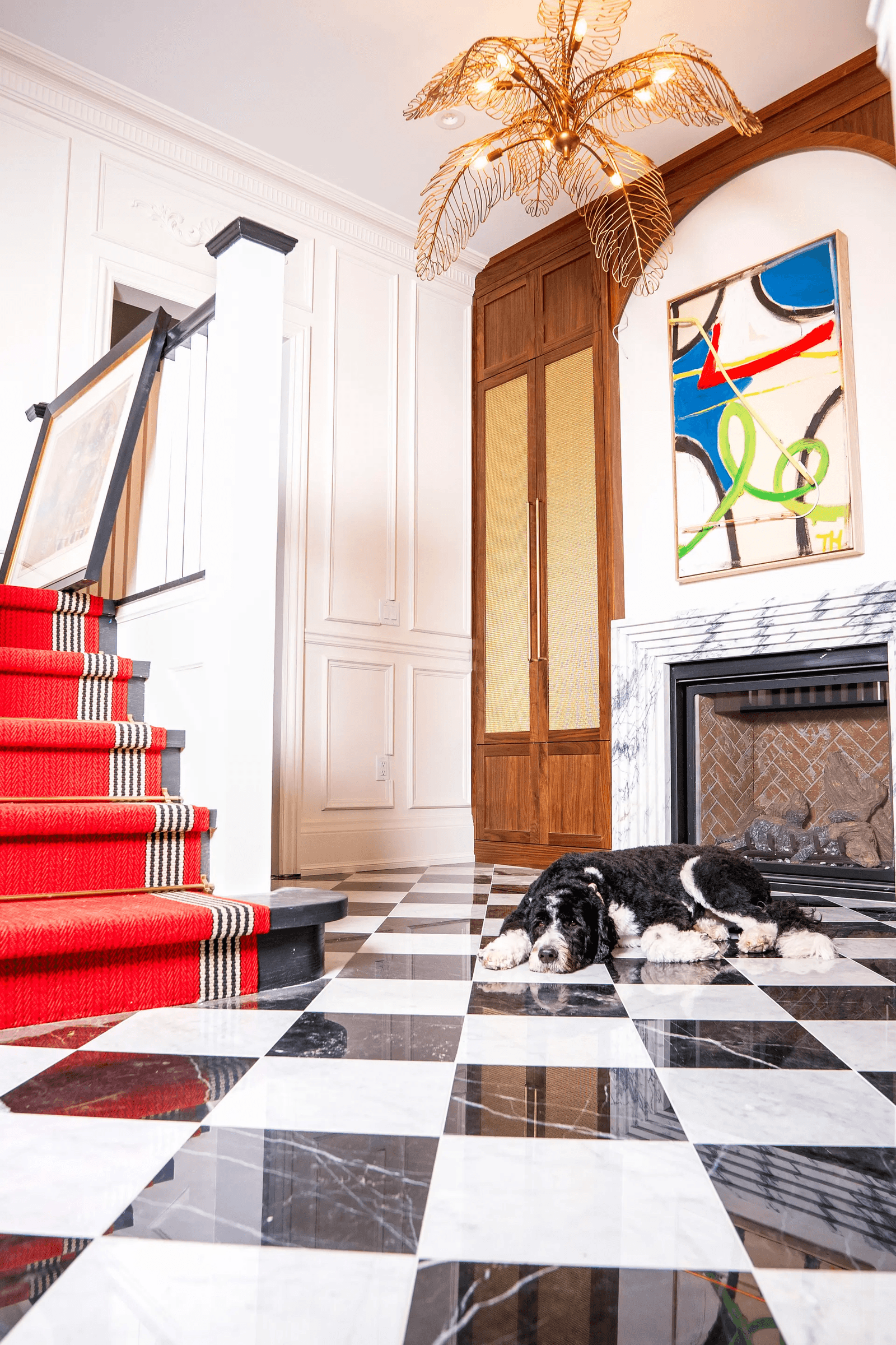 Elegant entryway with red carpeted staircase, marble fireplace, gold chandelier, and black and white checkered floor