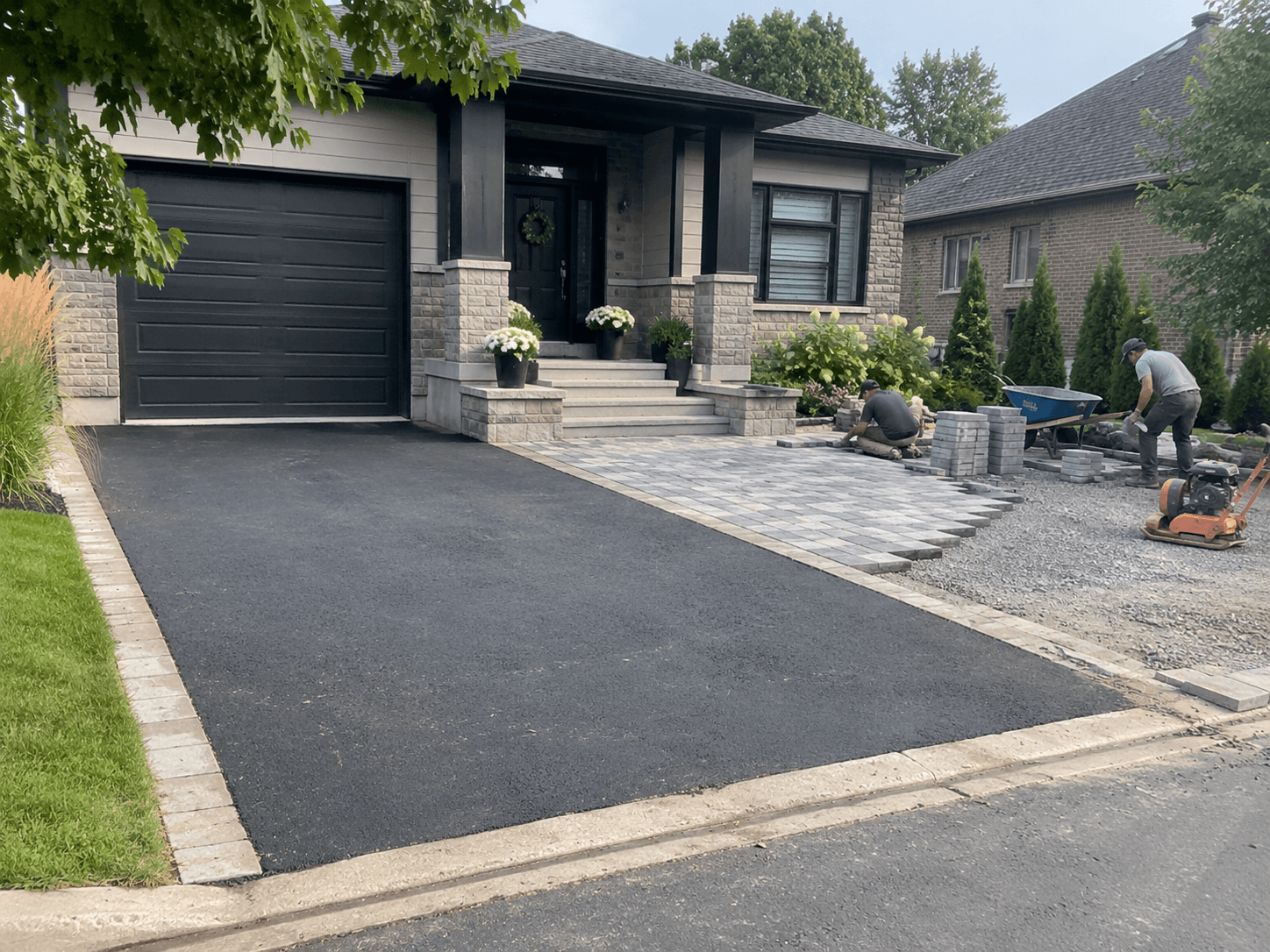 Workers installing grey interlocking pavers in front of a residential home with an asphalt driveway, paver edging, compacted gravel and landscaping