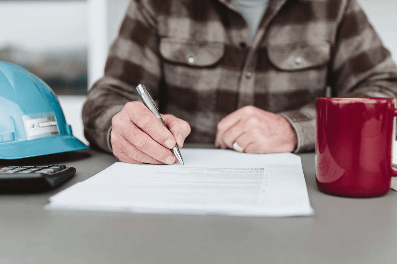 Construction professional filling out or signing a document at a table, with a hard hat, calculator and mug nearby