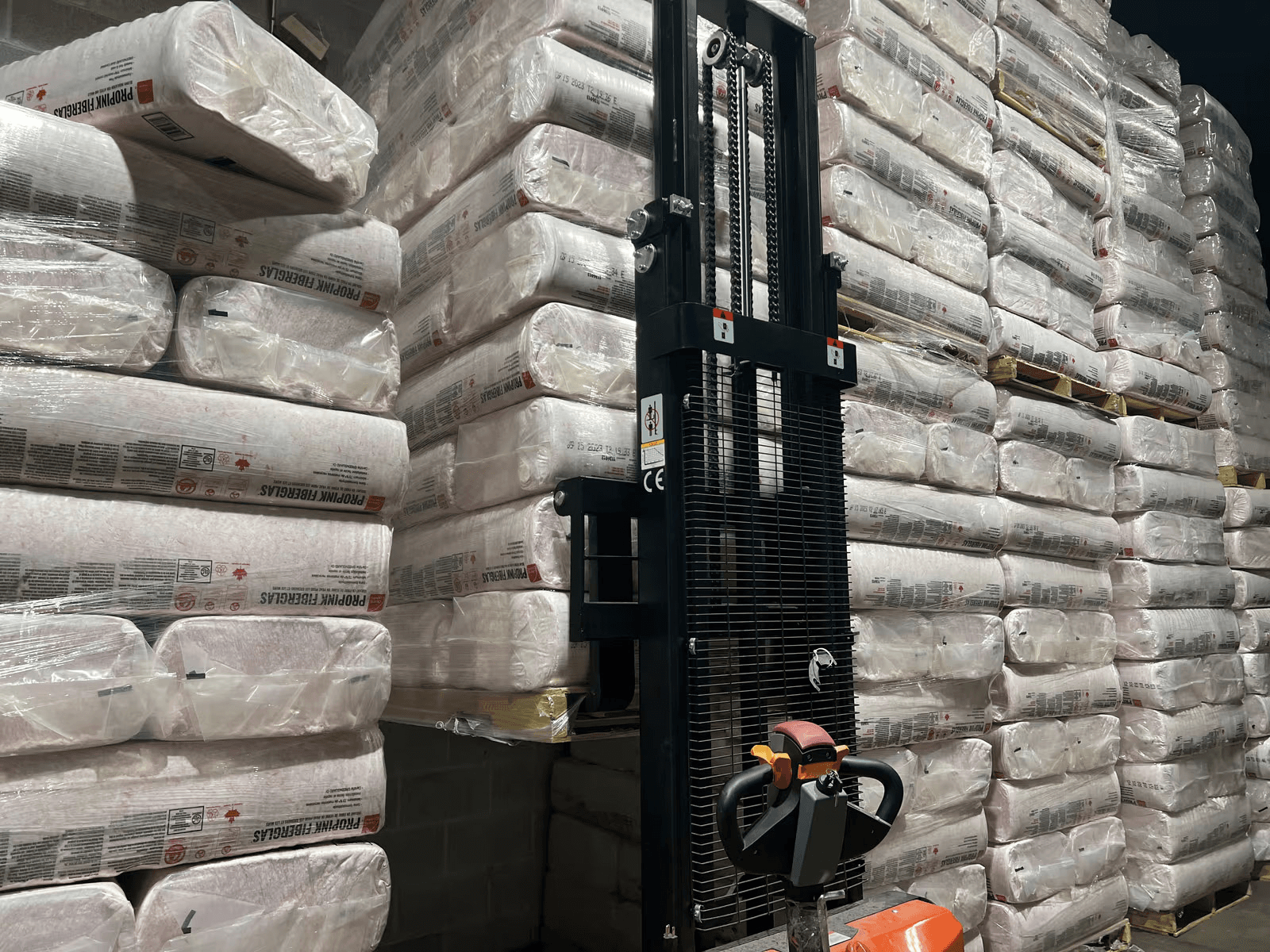 Warehouse filled with stacked fiberglass insulation packages on pallets, with an electric stacker used for material handling.