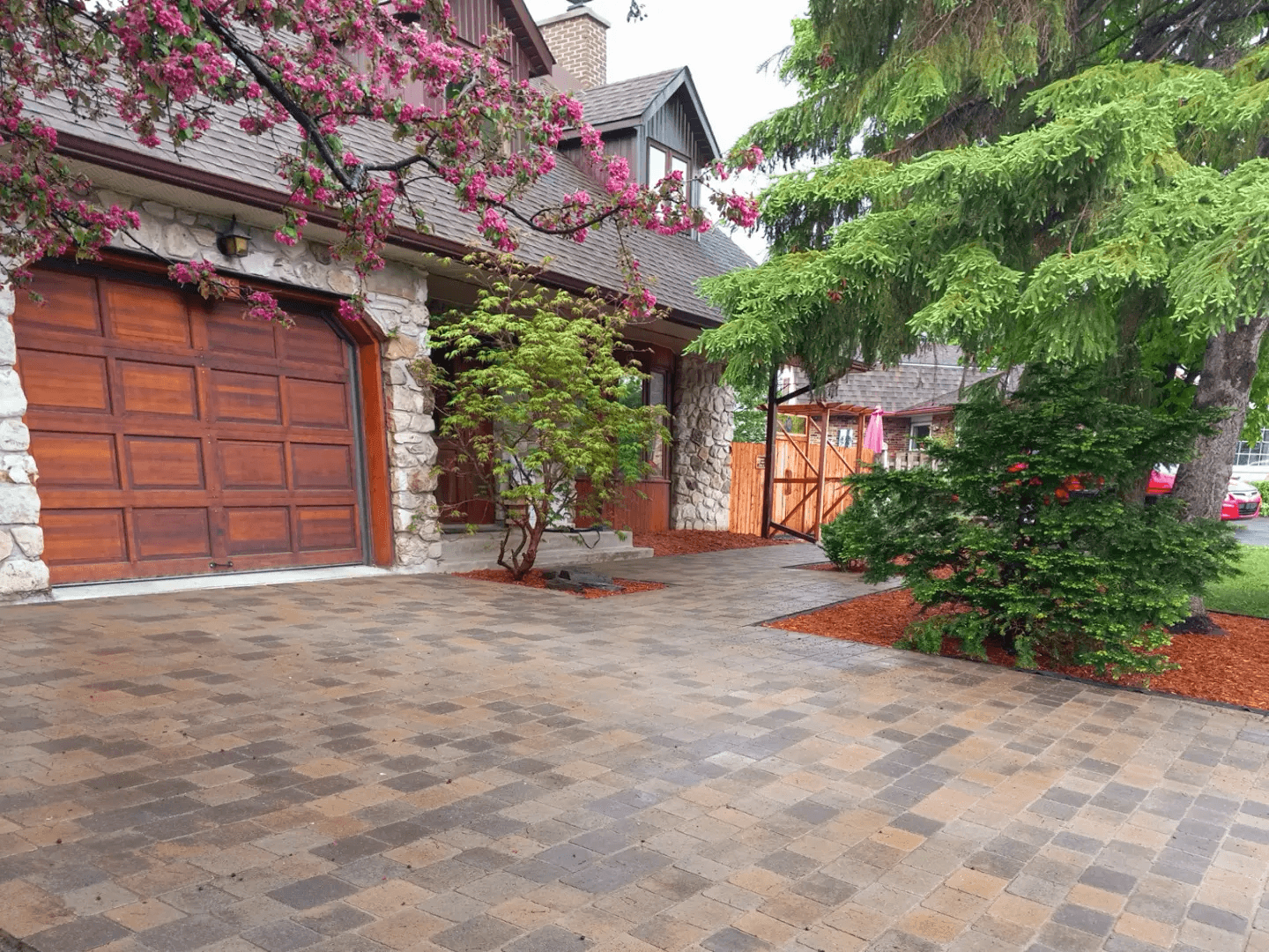 Multicolored interlocking paver driveway in front of a stone and wood home with a wooden garage door, mature trees and landscaped surroundings