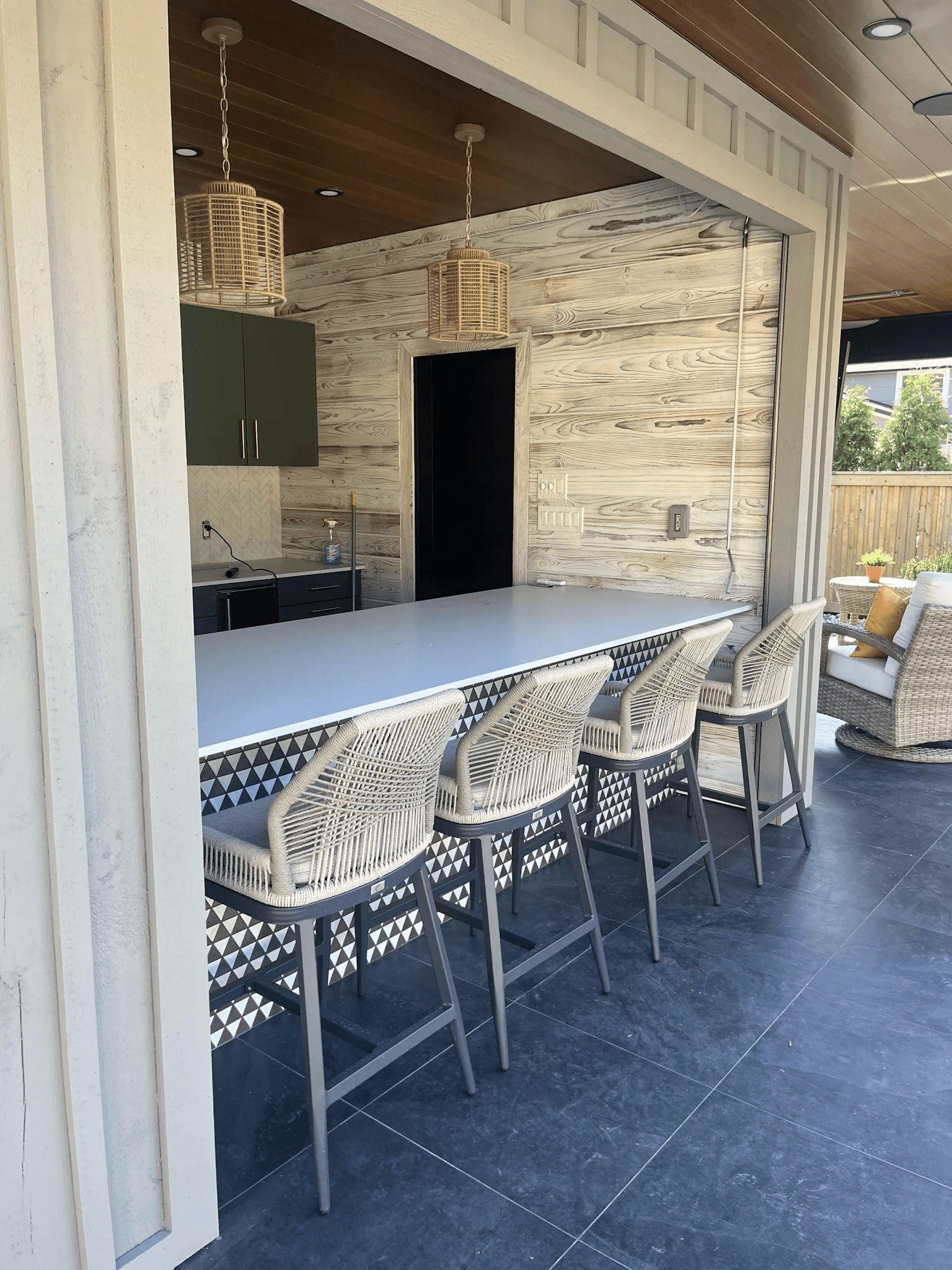 Covered outdoor kitchen with a bar counter, stools, built-in cabinets, pendant lighting and wood wall finishes.