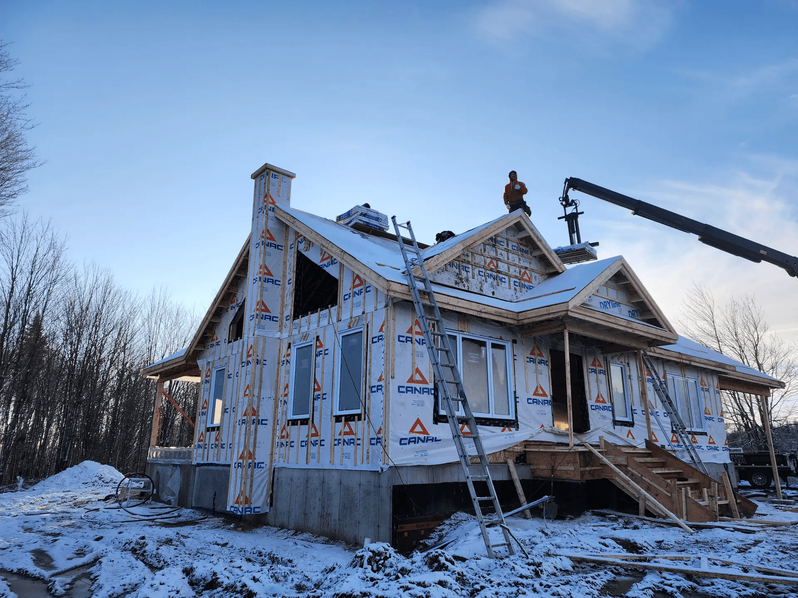 House under construction in winter with CANAC weather barrier wrap, scaffolding, ladders, and workers on the roof under a blue sky.