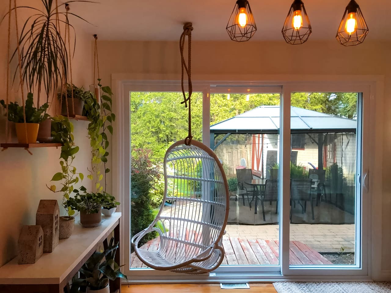 Bright relaxation corner with hanging chair, numerous indoor plants, pendant lights and large glass patio door overlooking a terrace with gazebo.