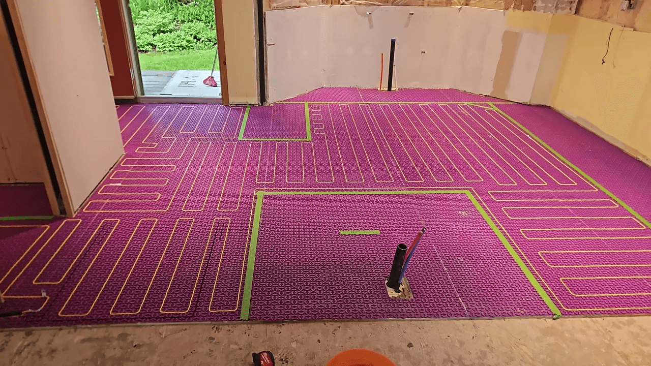 Hydronic radiant floor heating installation with purple uncoupling membrane, red tubing, green tape and plumbing rough-ins in a room under renovation