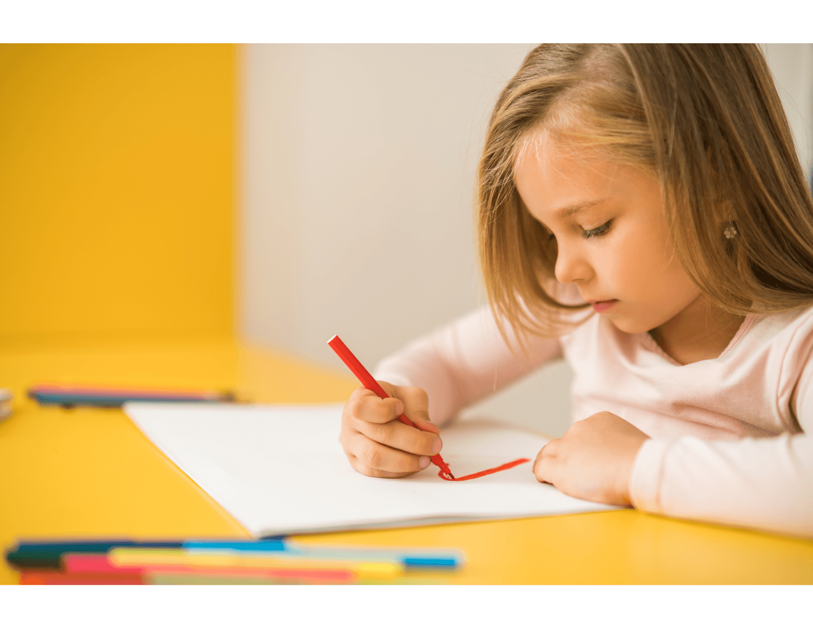  Little girl drawing with a red pencil on a sheet of paper, sitting at a yellow table.