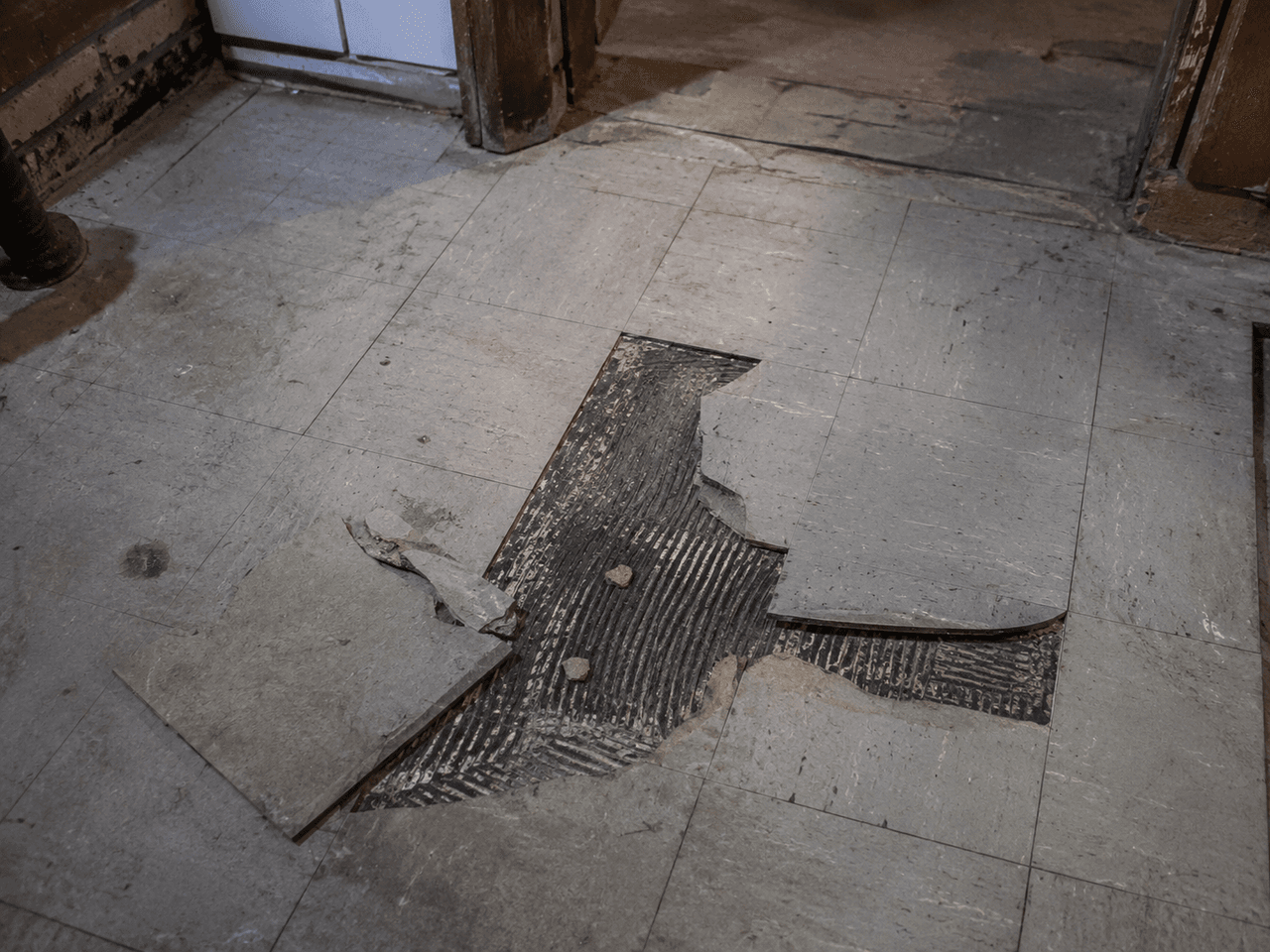 Damaged interior floor with removed tiles and exposed black adhesive, prepared for demolition and flooring replacement work