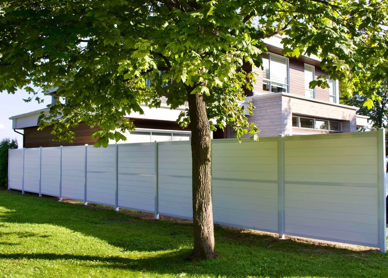 Modern white privacy fence installed in front of a contemporary home, surrounded by a green lawn and a large mature tree.