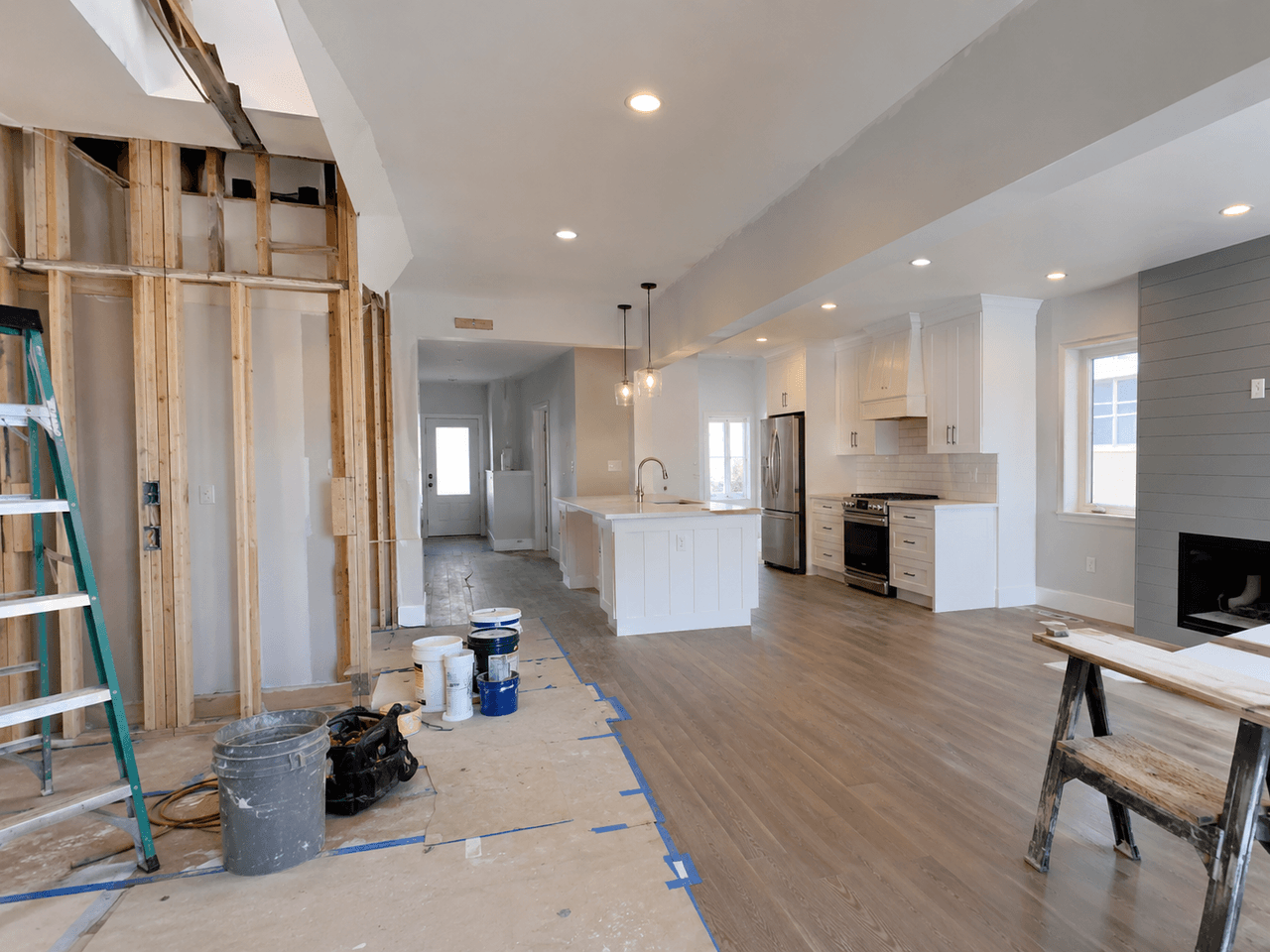 Open-concept interior under renovation with a white kitchen, central island, wood flooring, modern fireplace, and a partially opened wall exposing the framing.