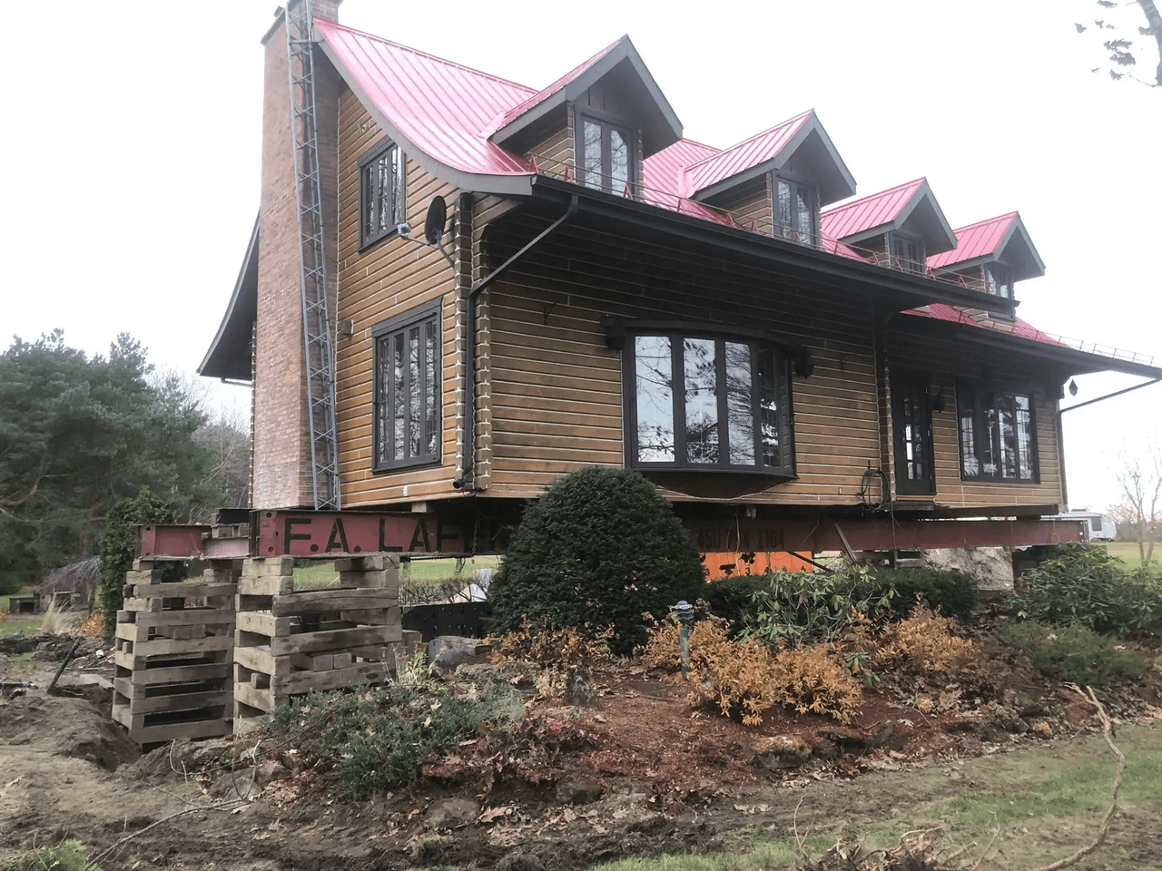 Elevated wooden house with red metal roof, brick chimney, black windows and landscaping under construction