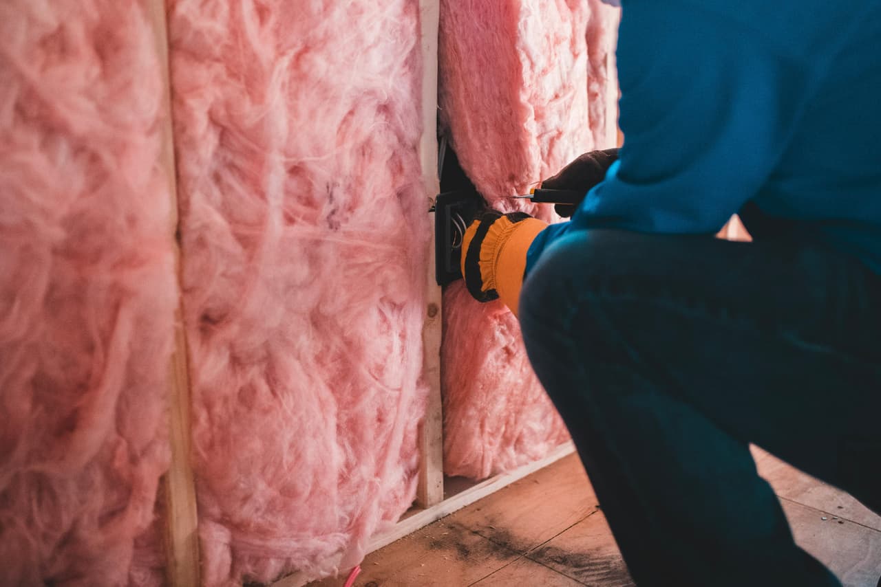 Worker performing an electrical installation inside a residential wall insulated with pink fiberglass during construction or renovation work.